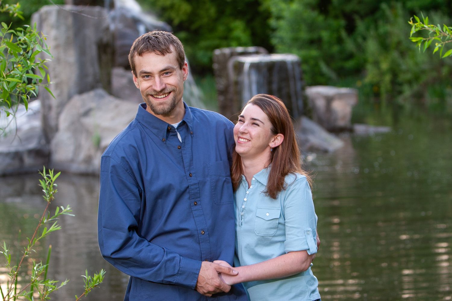 Smiling couple holding hands near a waterfall surrounded by green trees.