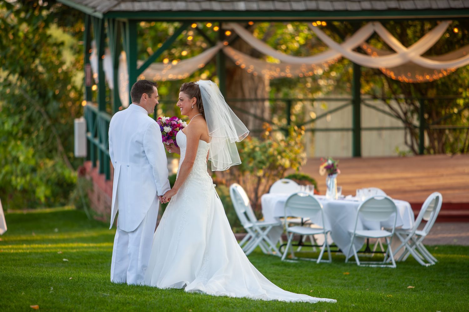A bride and groom stand face to face on a green lawn beneath a decorated gazebo during their outdoor wedding reception.