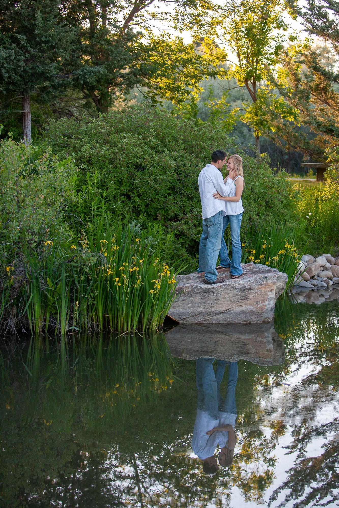 Couple standing on a rock kissing by a serene pond with lush green foliage and their reflection in the water.