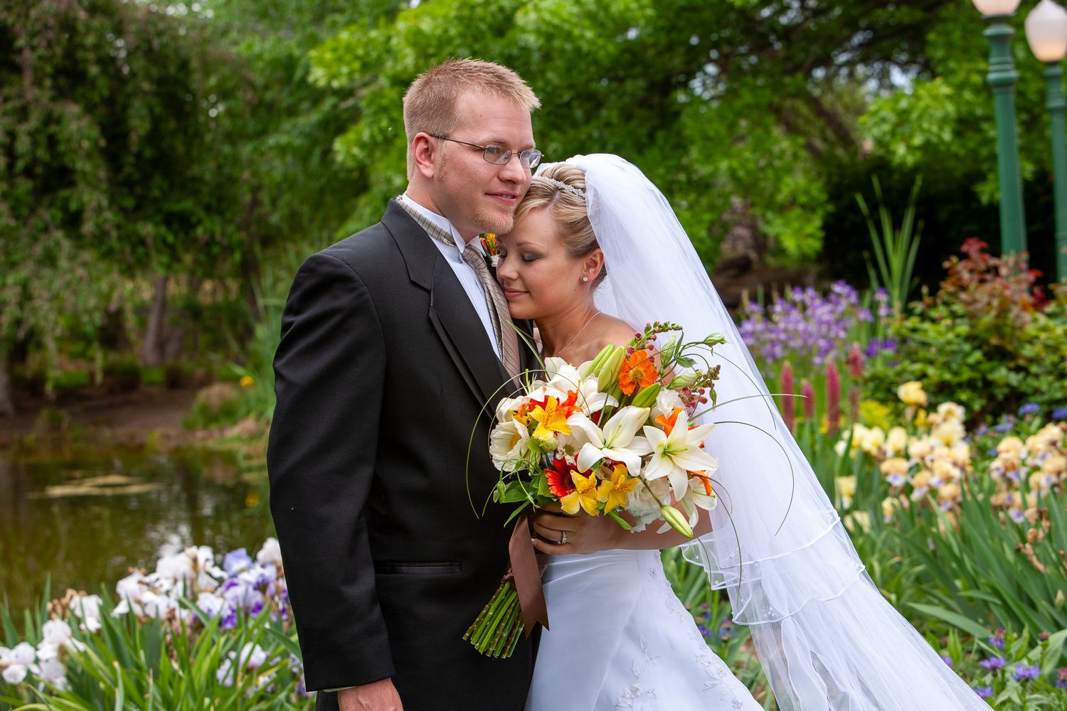 Bride and groom embracing in colorful garden with orange and white floral bouquet.