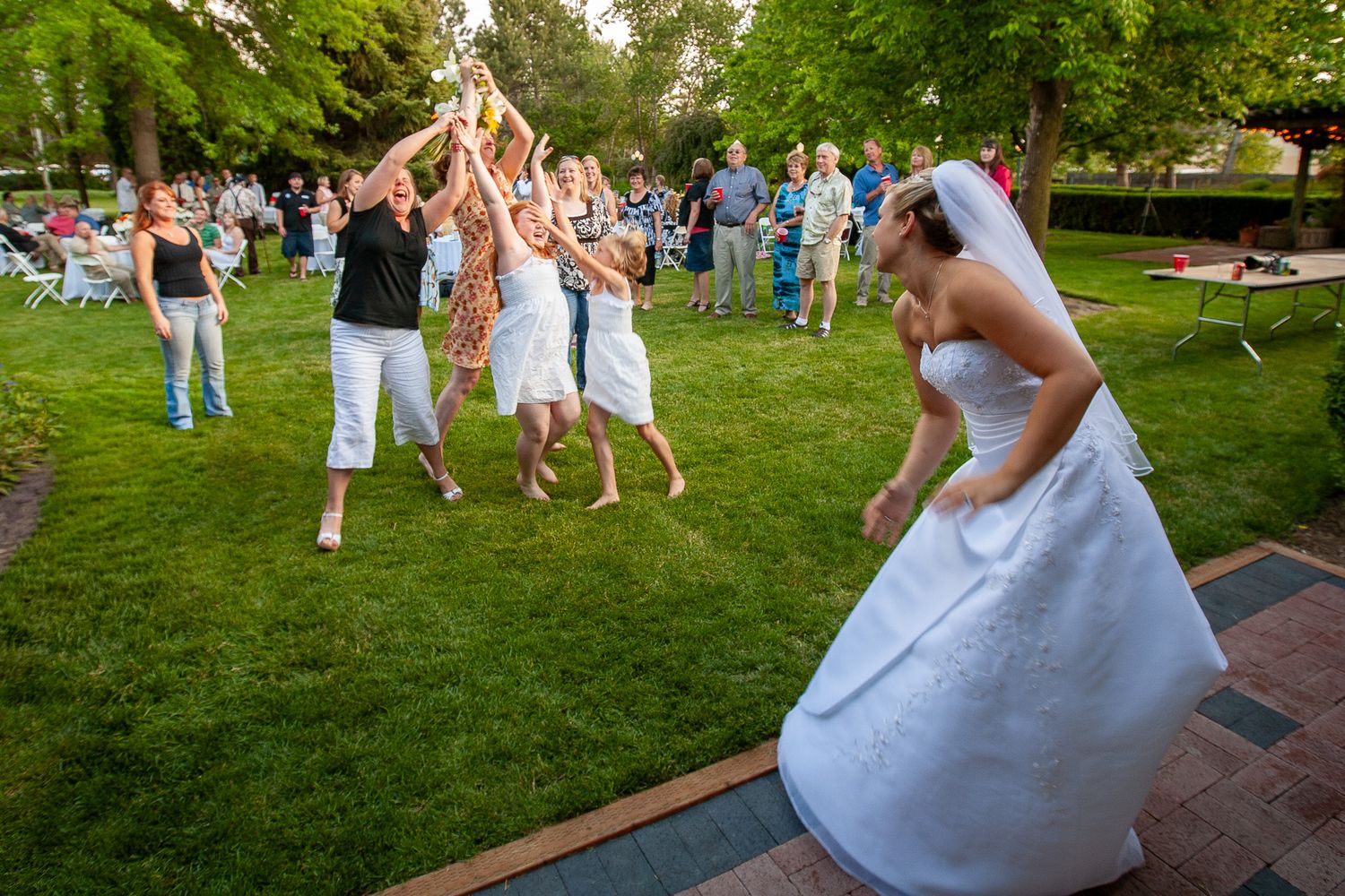 Bride in white gown tossing bouquet to excited guests on green lawn at outdoor wedding reception.