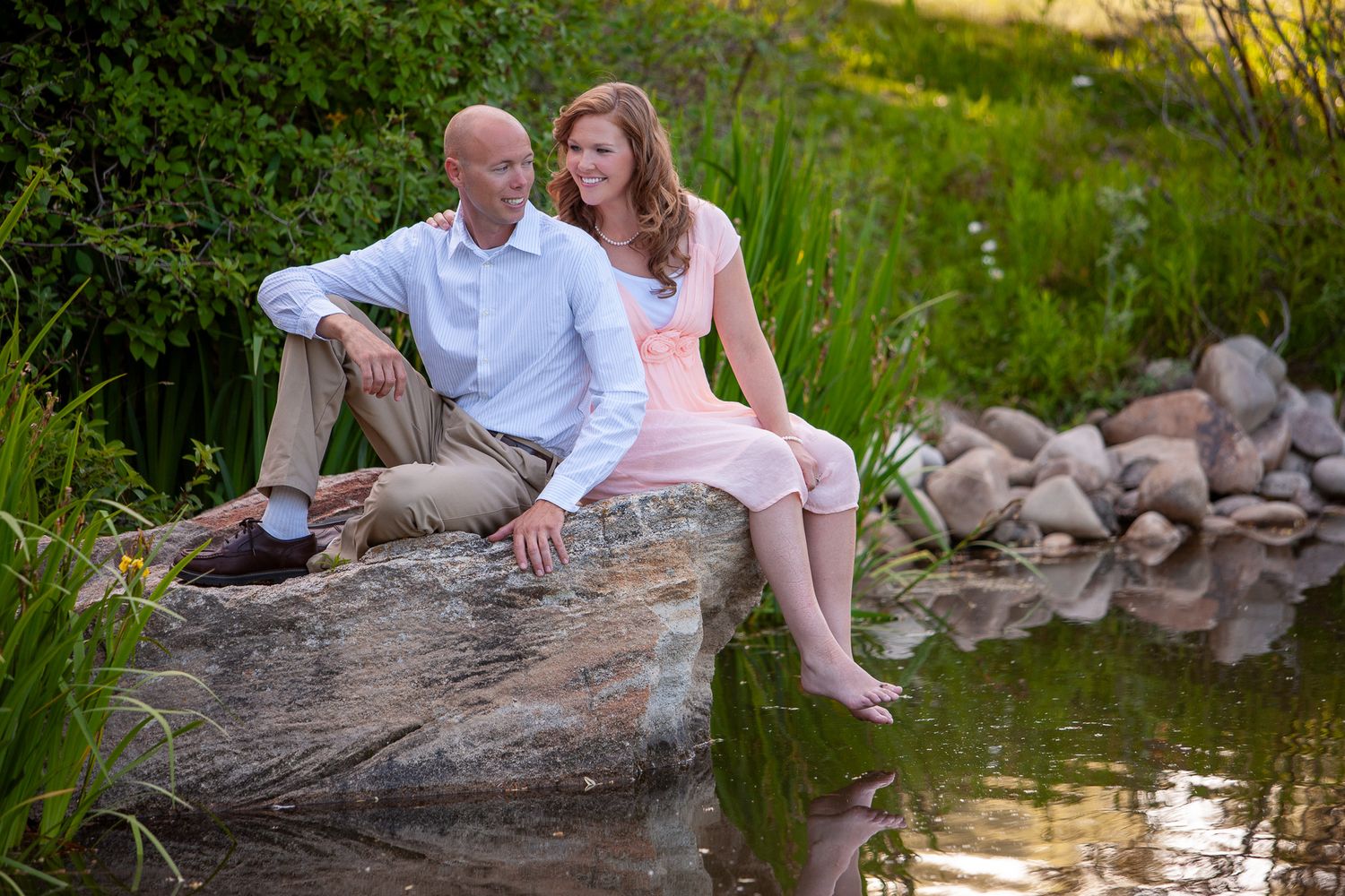 Romantic couple relaxes on rock by tranquil pond with green foliage and stone backdrop.
