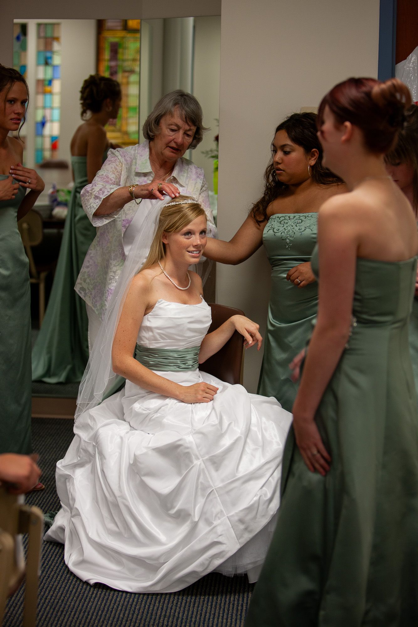 Bride seated in white gown surrounded by bridesmaids in sage green dresses getting ready.