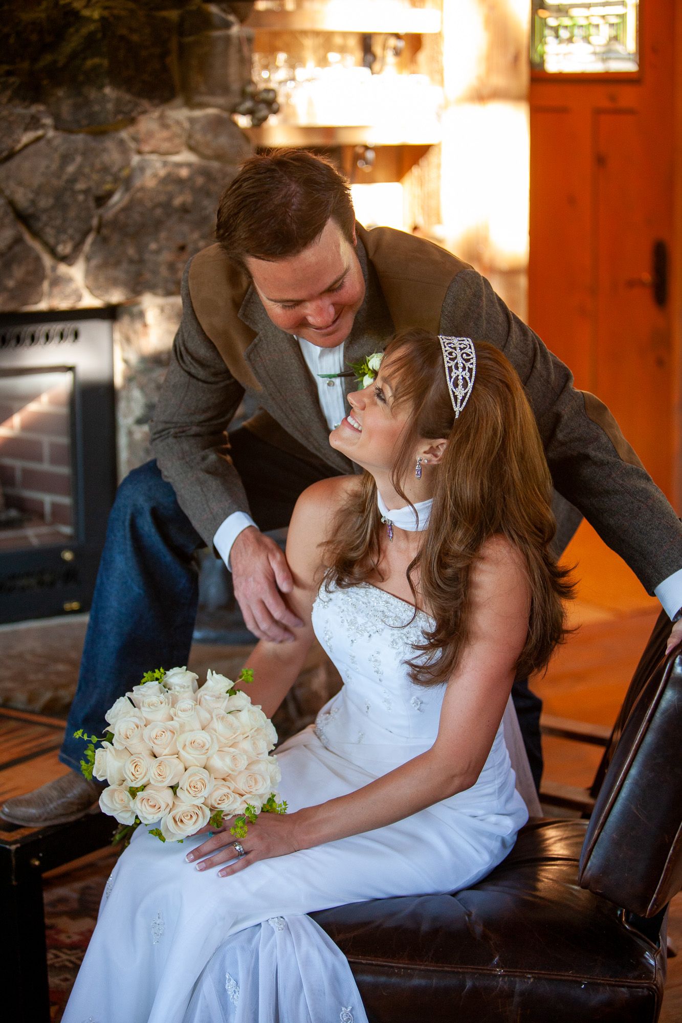 A groom in a tweed jacket leaning over his seated bride holding white roses near a stone fireplace.