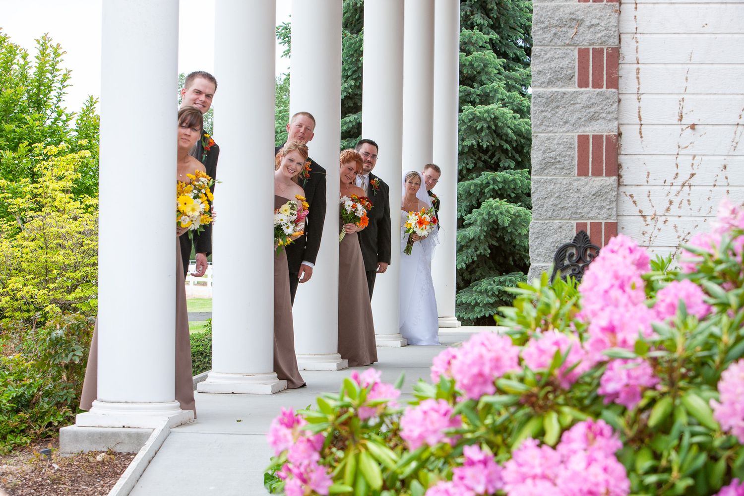 Wedding party peeking from behind white columns surrounded by pink flowering bushes.