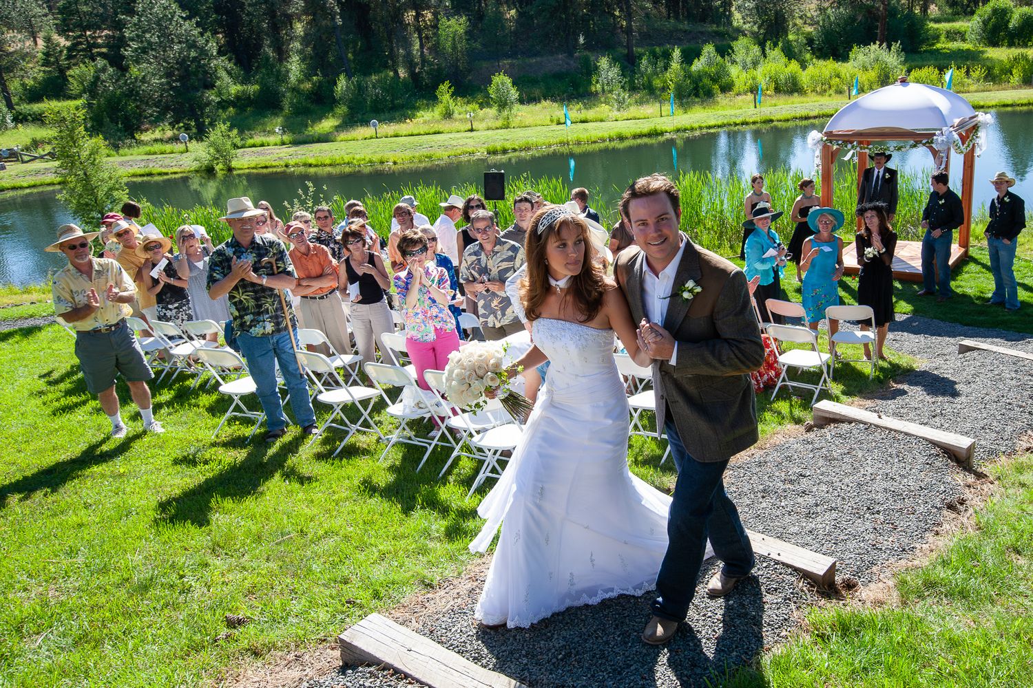Bride and groom walk down the aisle at an outdoor wedding ceremony by a scenic pond with guests seated on white chairs.