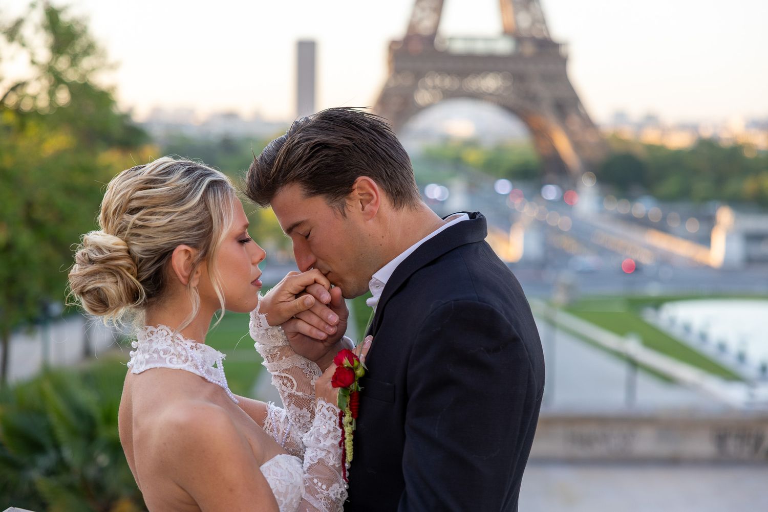 Groom kissing bride's hand tenderly with the Eiffel Tower glowing at sunset behind them.