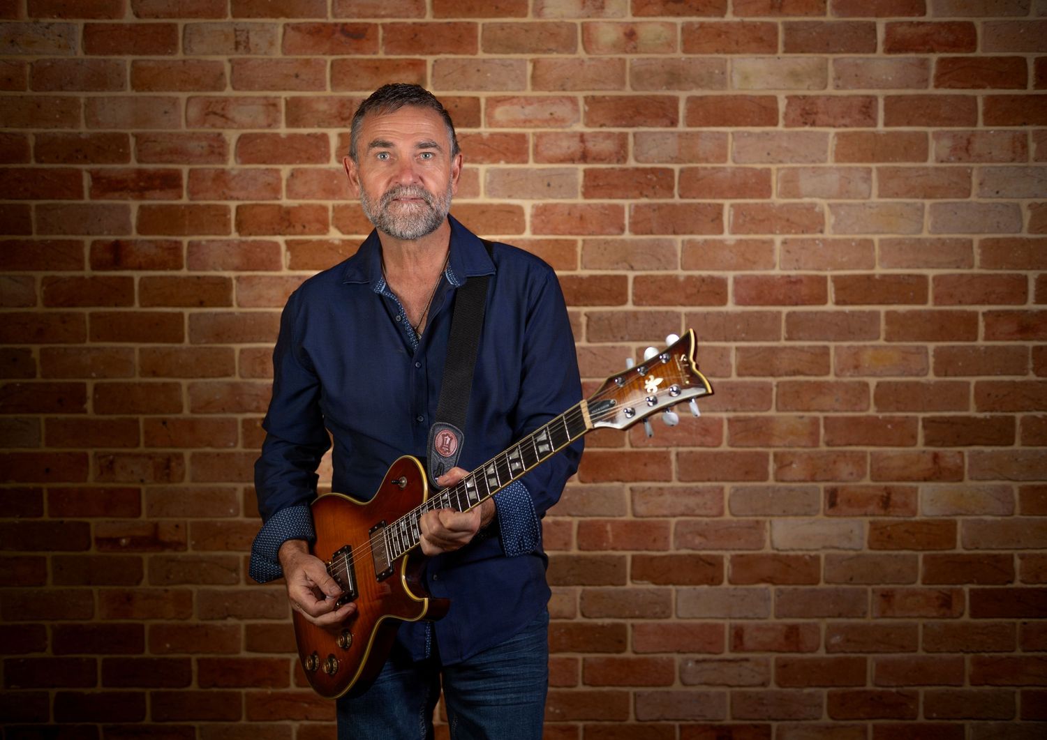 Middle-aged man in dark blue shirt playing a semi-hollow electric guitar against a red brick wall.