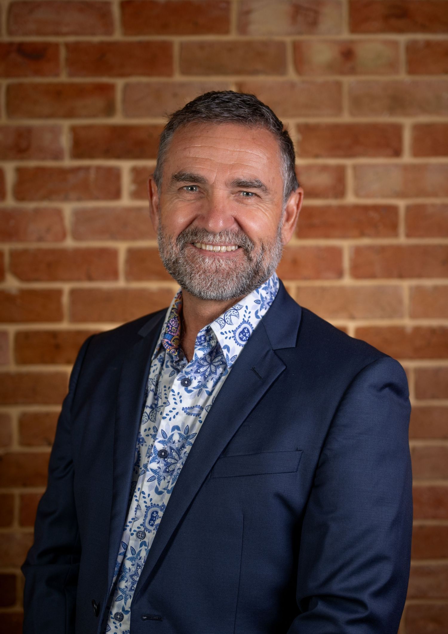Professional man in navy blazer and floral shirt smiling confidently against a brick wall background.