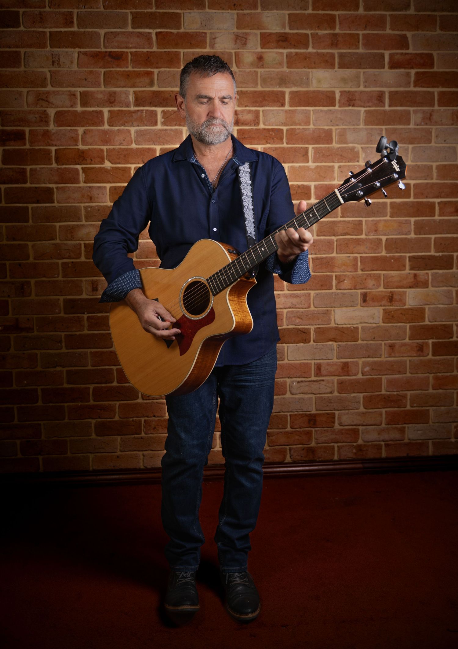 Middle-aged man playing acoustic guitar while standing against a red brick wall indoors.