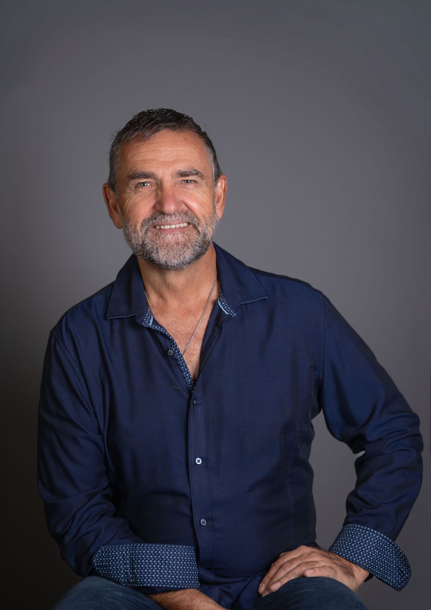Middle-aged man with gray beard smiling, wearing a navy blue button-up shirt, posed against a dark gray background.