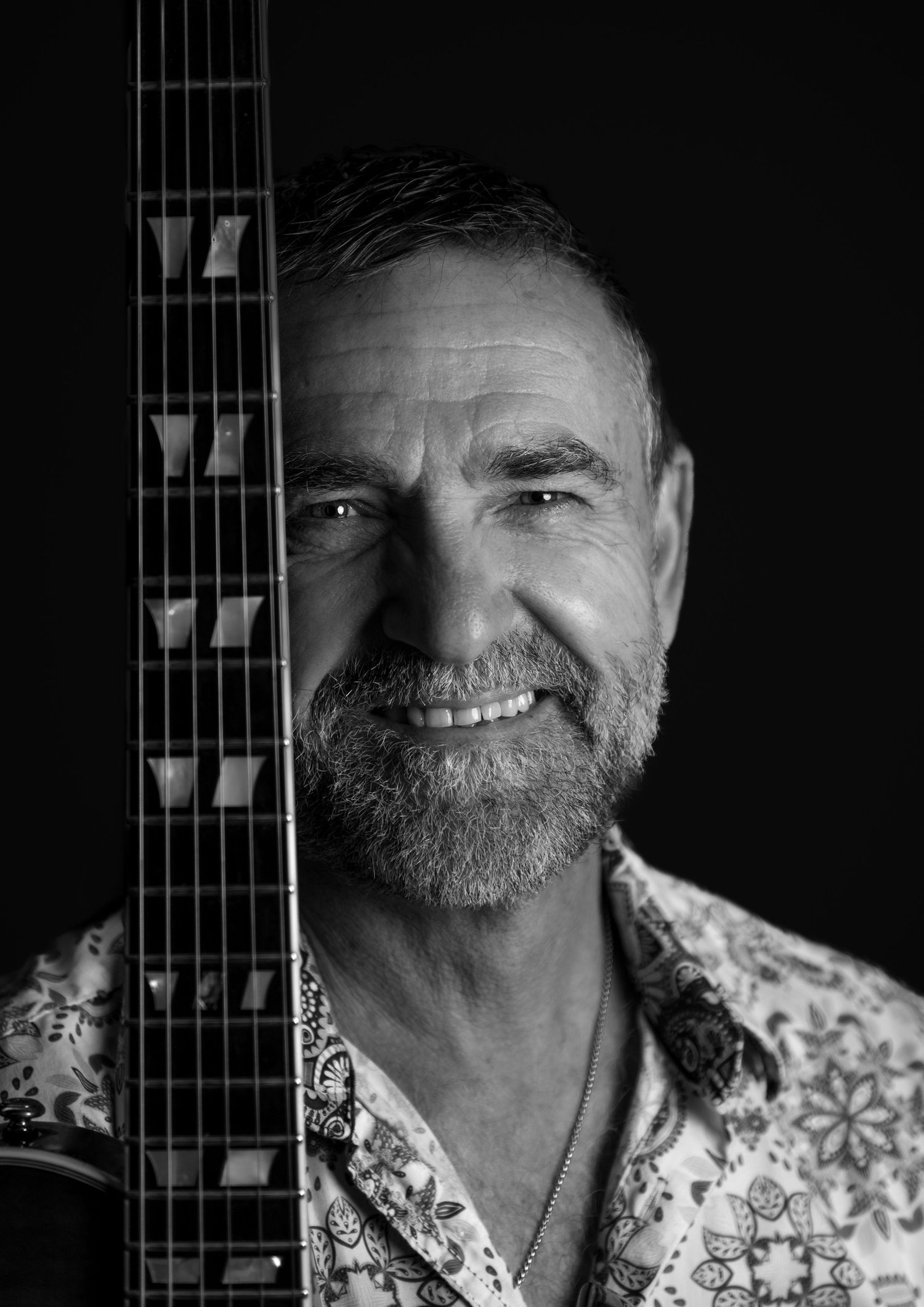 Black and white portrait of a bearded man smiling beside an acoustic guitar neck in dramatic lighting.