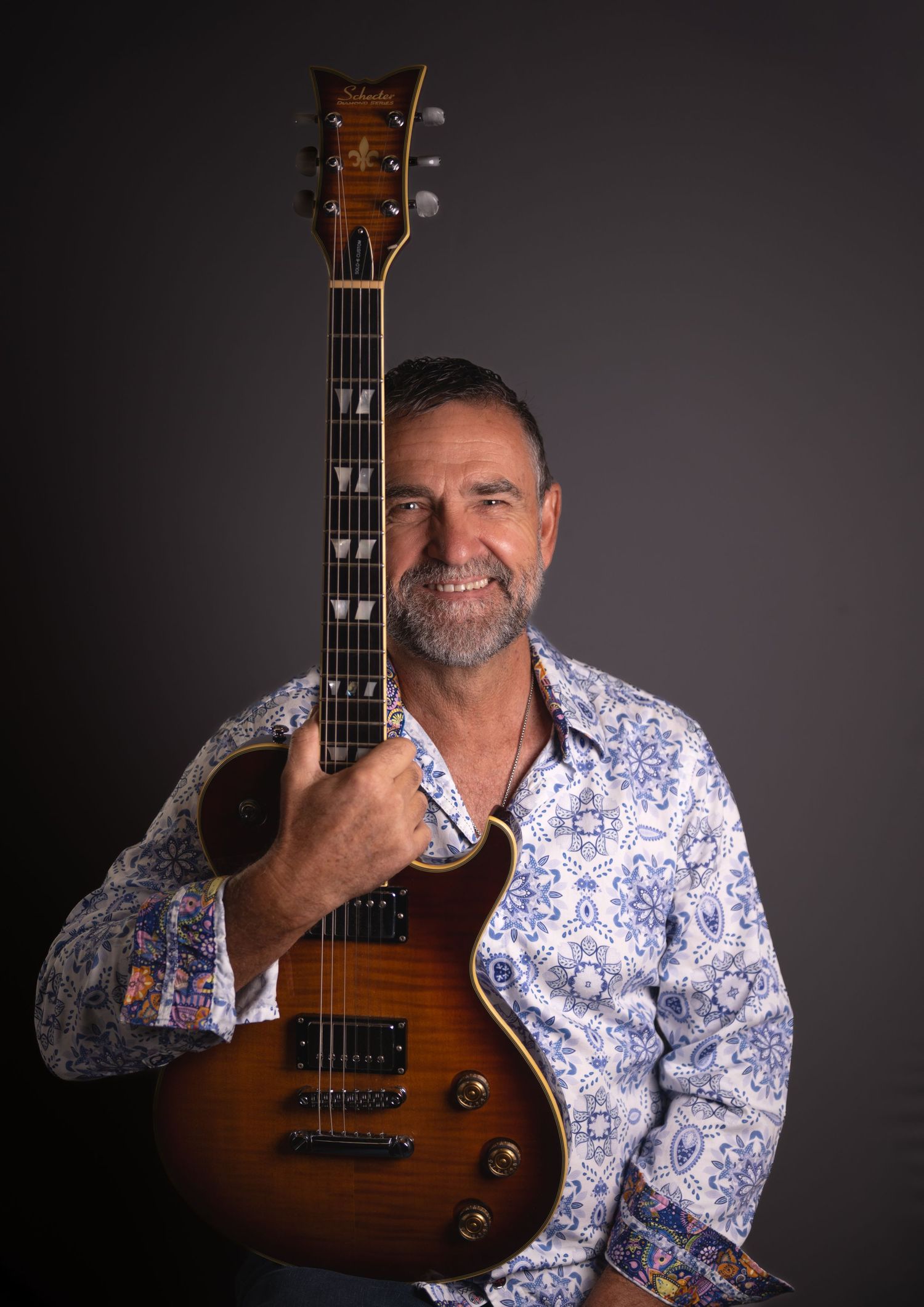 Man in floral shirt smiling while holding a sunburst electric guitar against a dark studio background.