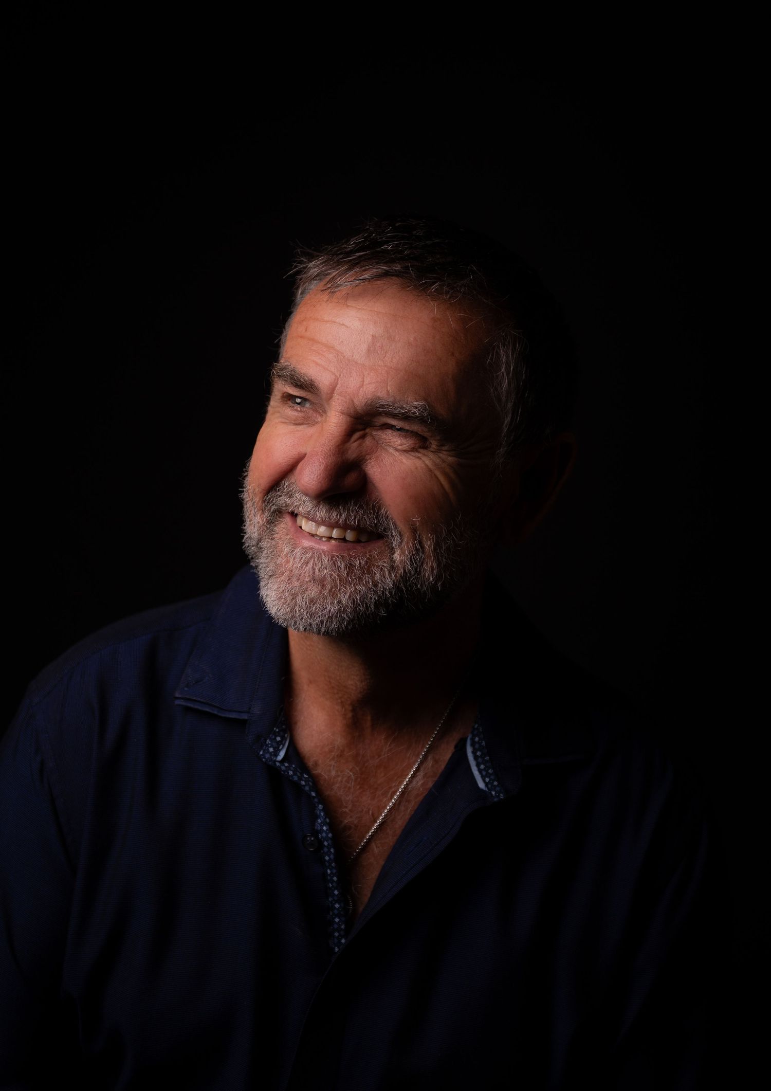 Smiling middle-aged man with gray beard wearing dark blue shirt, portrait against black background.