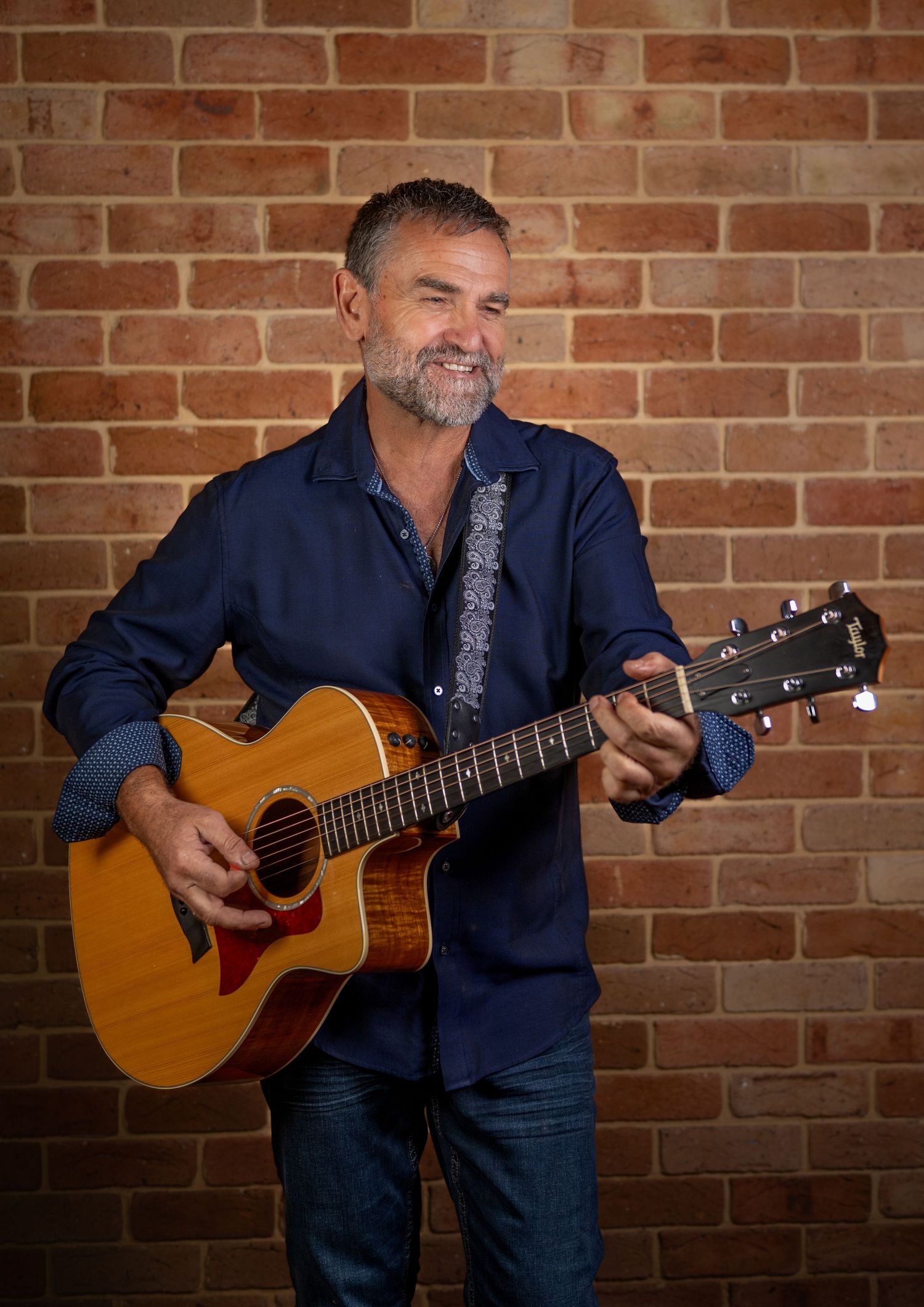 Smiling middle-aged man playing acoustic guitar against a brick wall, wearing a navy blue shirt.