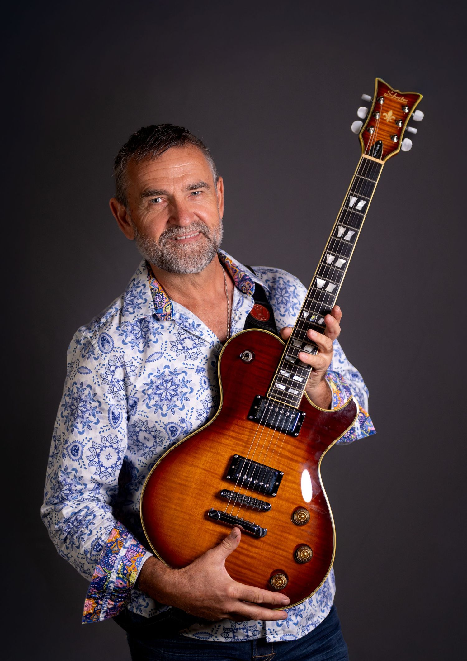 Middle-aged man with beard holding a sunburst Les Paul electric guitar, wearing a floral shirt against dark background.
