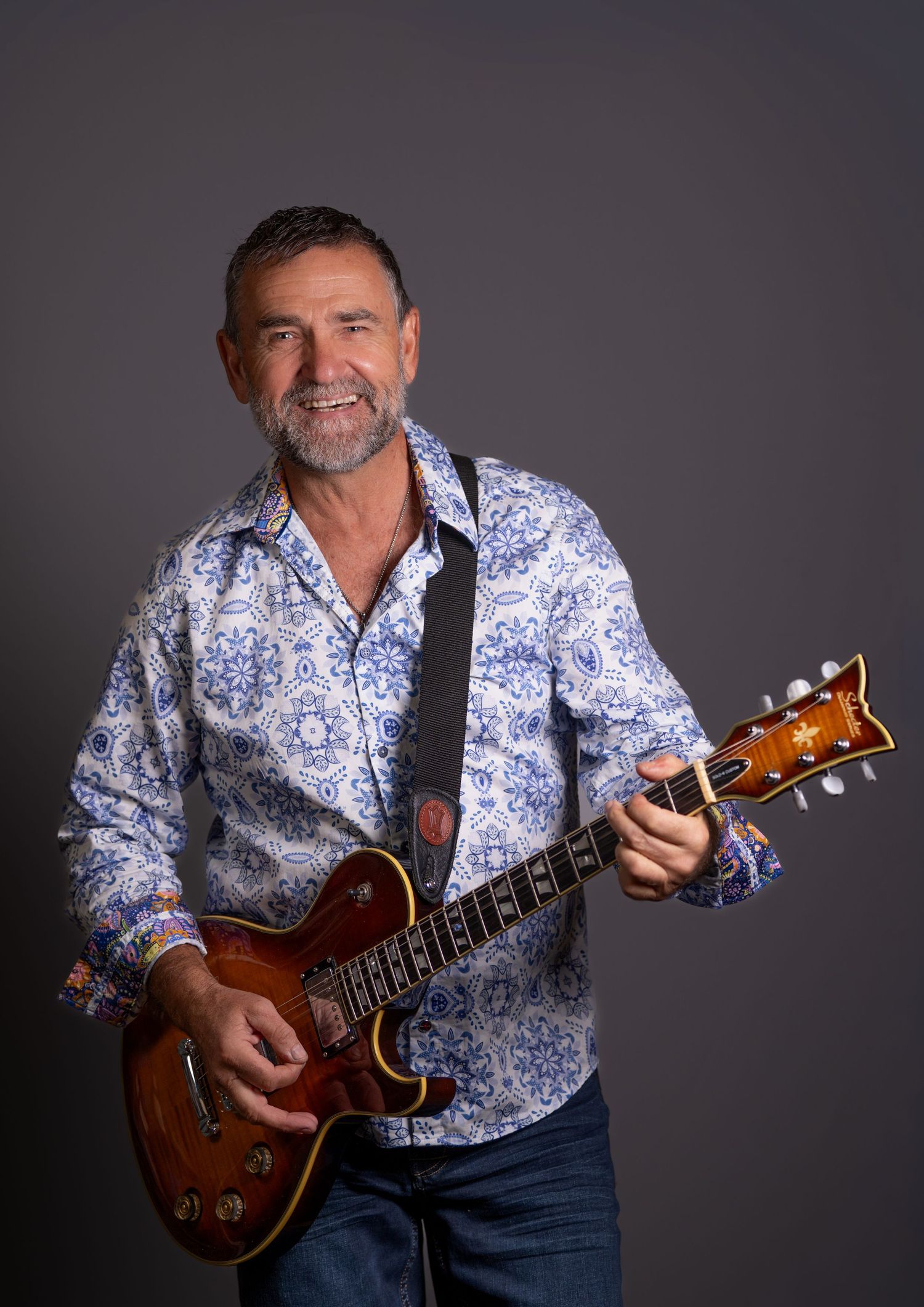 Middle-aged man smiling while playing a brown semi-hollow electric guitar, wearing a blue floral shirt.