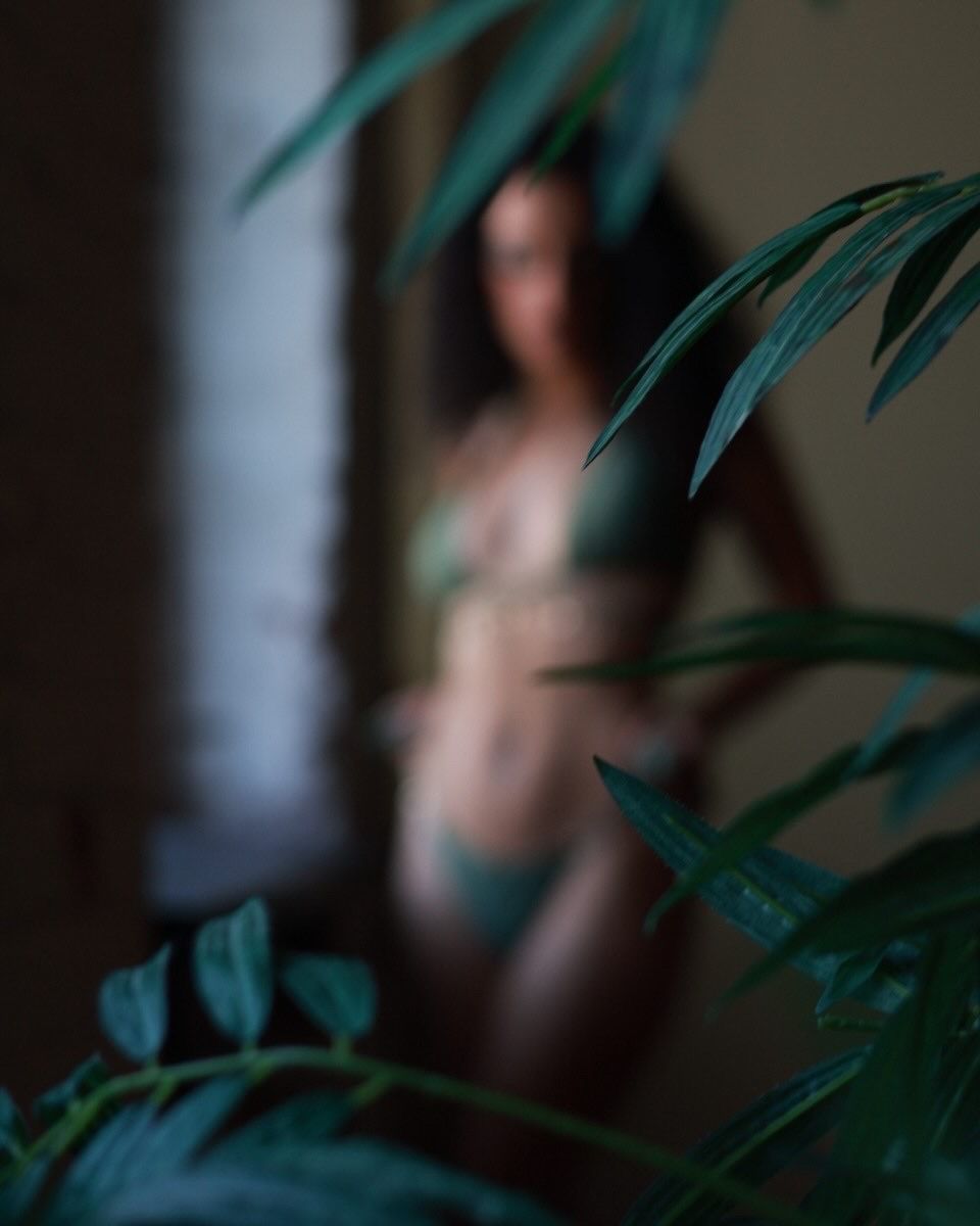 A woman in green lingerie stands behind lush tropical leaves in a moody, softly lit indoor setting.