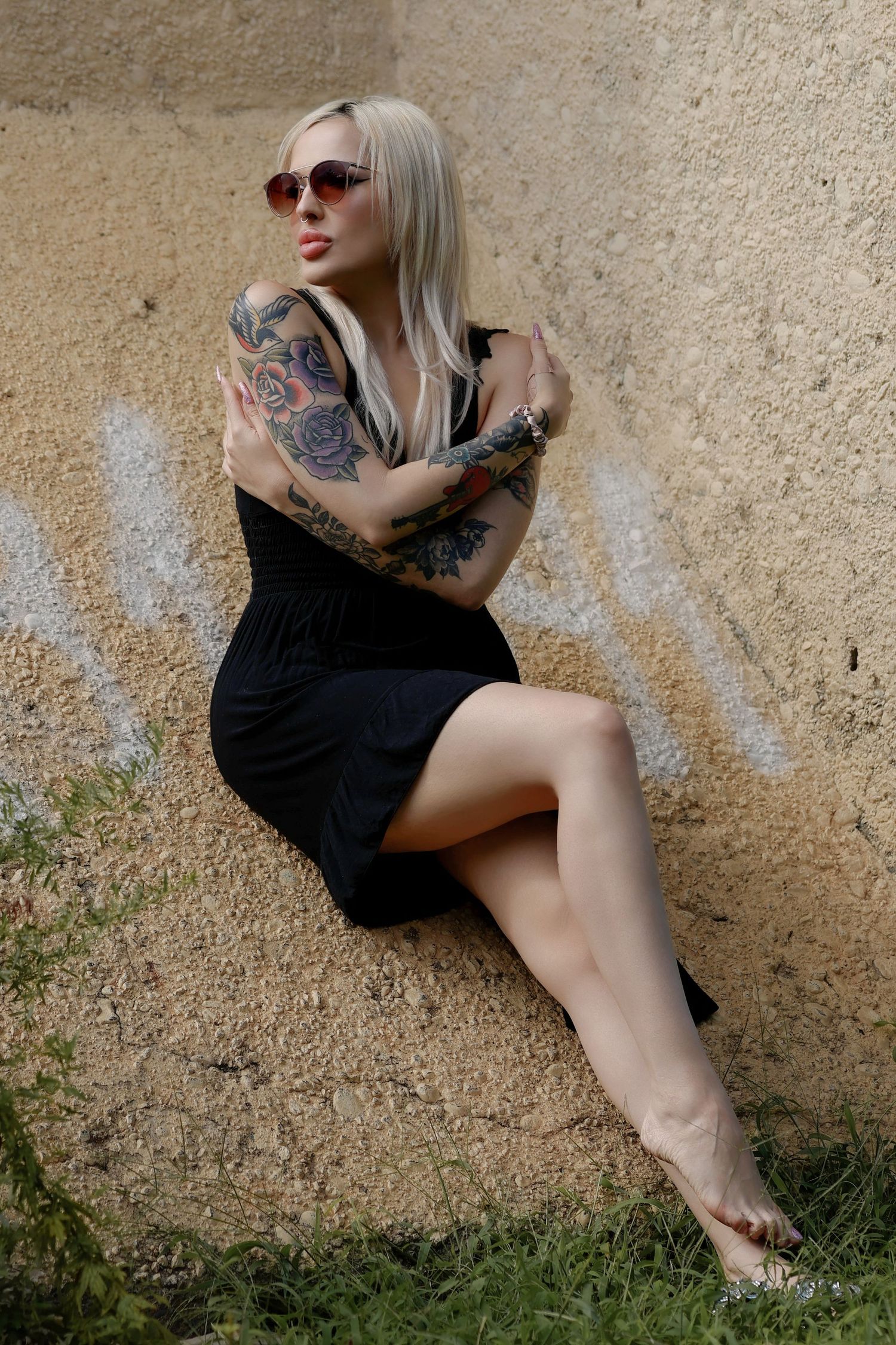 Tattooed blonde woman in black dress and sunglasses sitting barefoot against a textured wall outdoors.