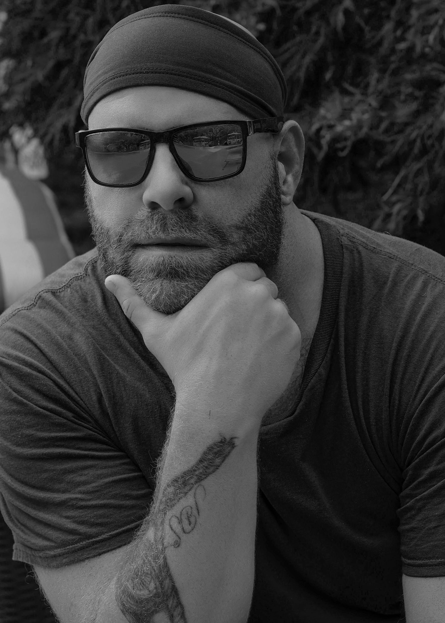 Black and white portrait of bearded man wearing bandana and sunglasses with tattoos, resting chin on hand.