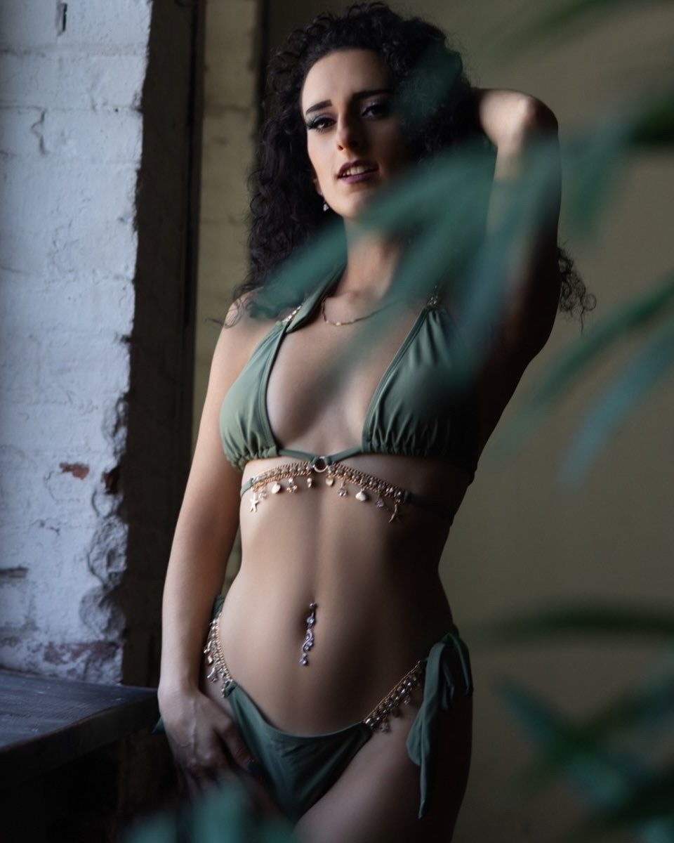 Woman in green bikini with gold chain belt posing gracefully among tropical leaves in soft natural light.