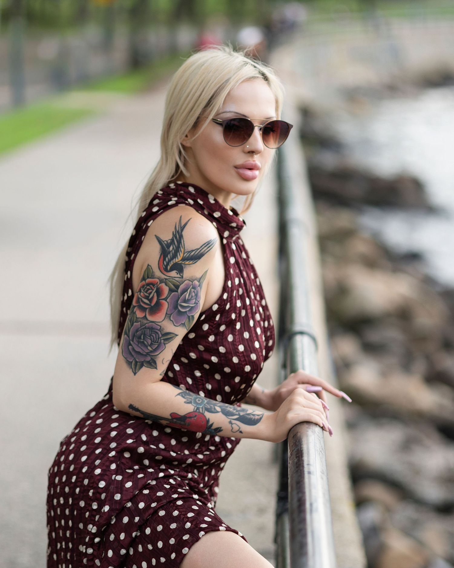 Blonde woman with tattoos wearing a polka dot dress and sunglasses leaning on a railing by the waterfront.