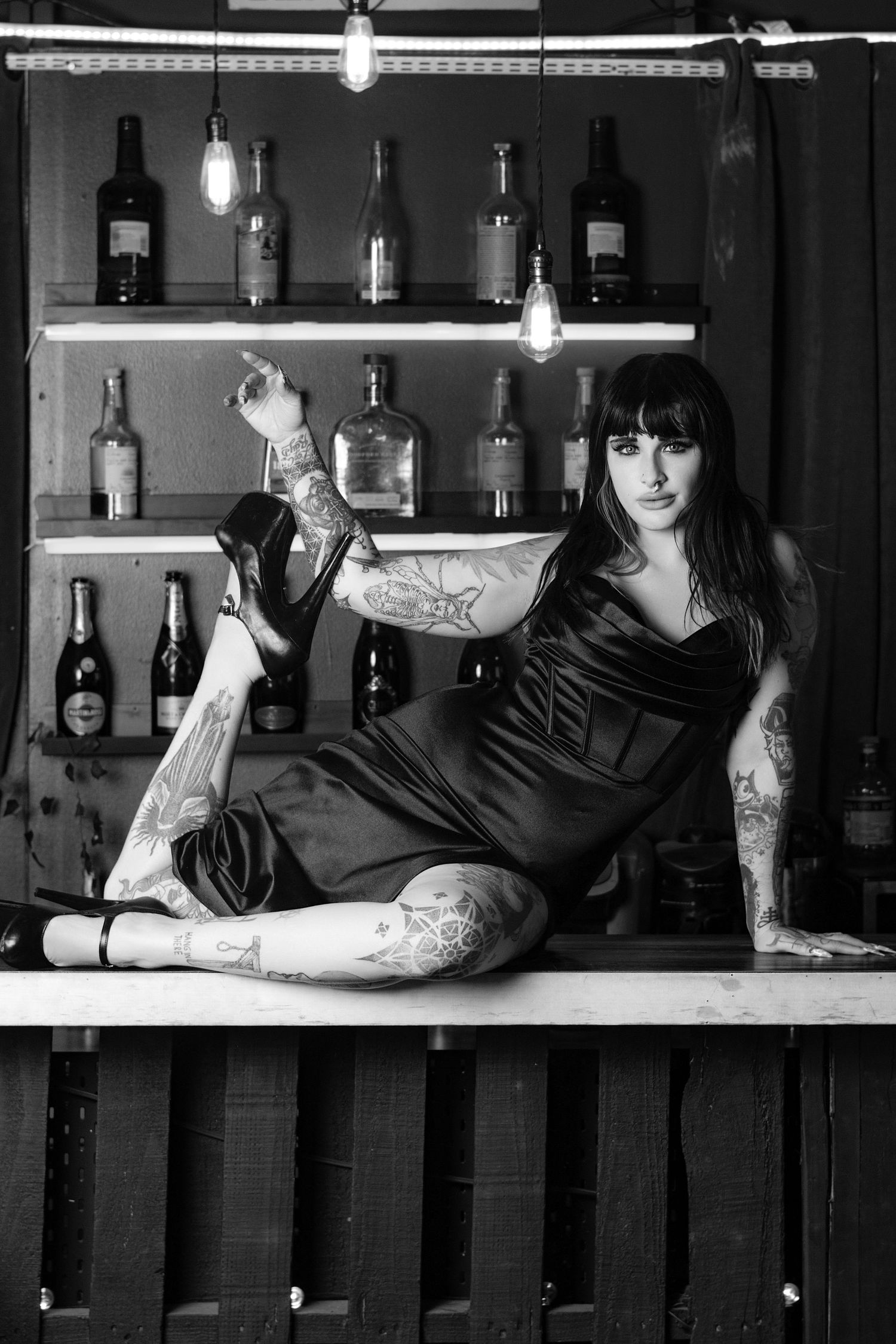 Black and white photo of tattooed woman in black dress posing on bar counter with liquor bottles on shelves behind her.