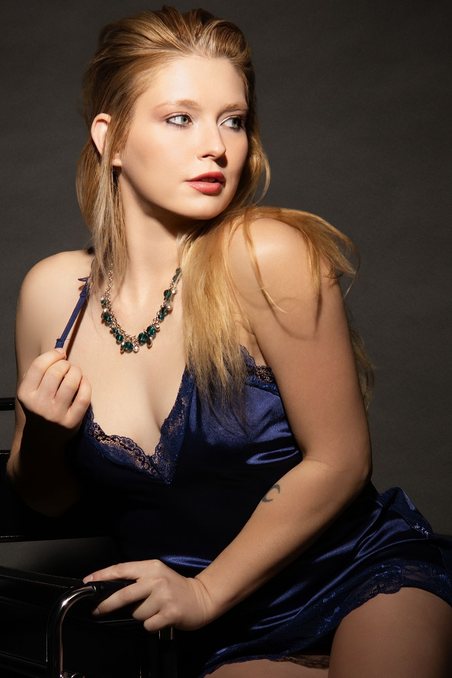 Blonde woman in blue satin slip dress with green gemstone necklace poses elegantly in a studio setting.
