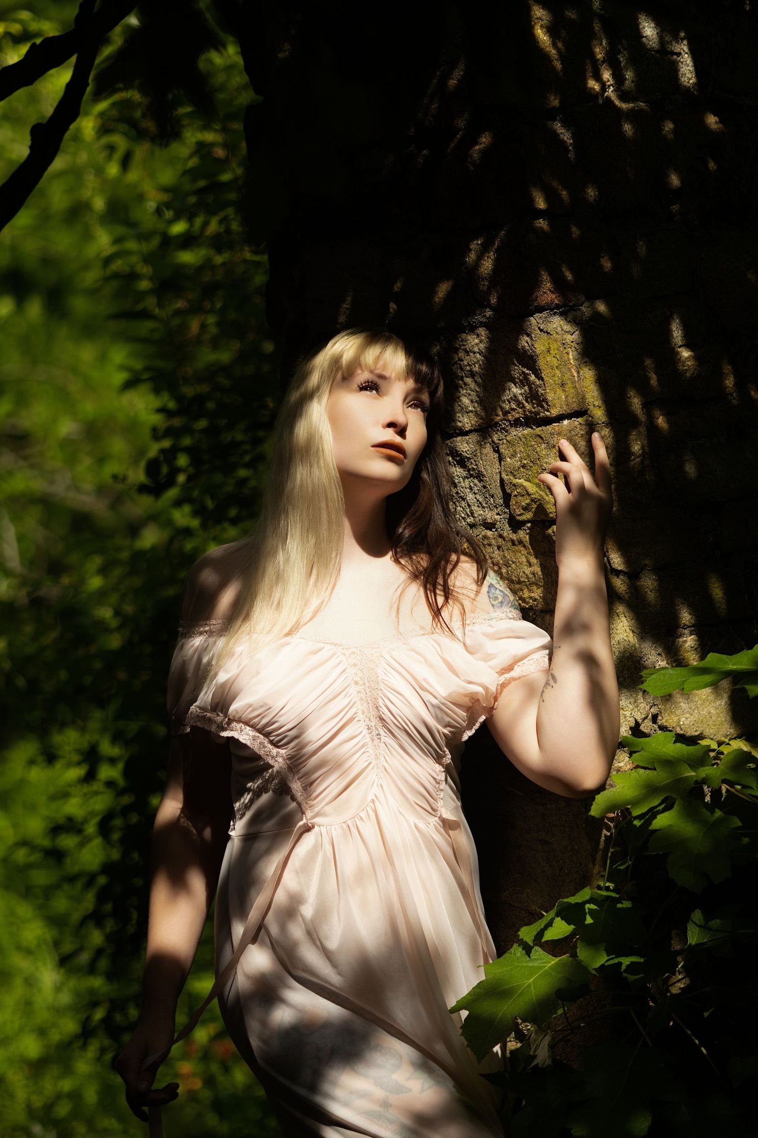 Blonde woman in pink dress leaning against mossy tree in sunlit forest.