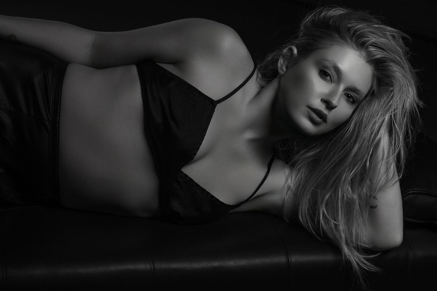 Black and white photo of a blonde woman in a dark camisole reclining on a leather sofa.