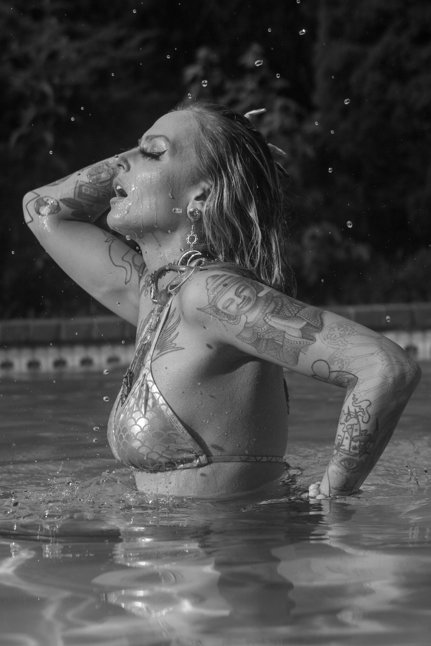 Black and white photo of tattooed woman in bikini emerging from pool with water droplets falling around her.