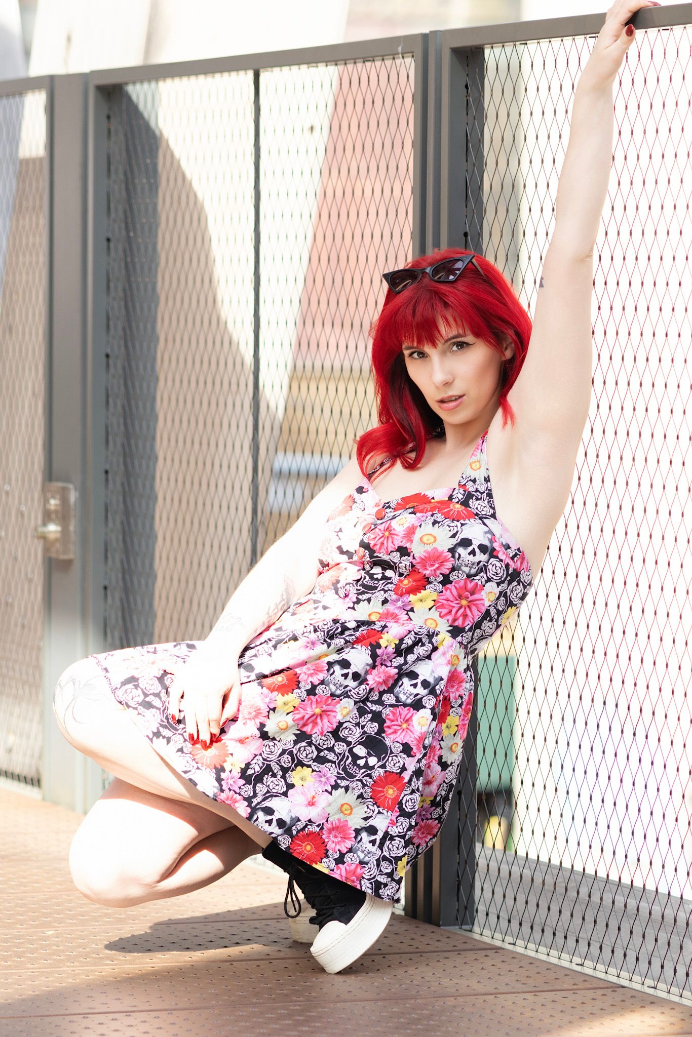 Red-haired woman in floral skull dress crouching by metal fence outdoors.