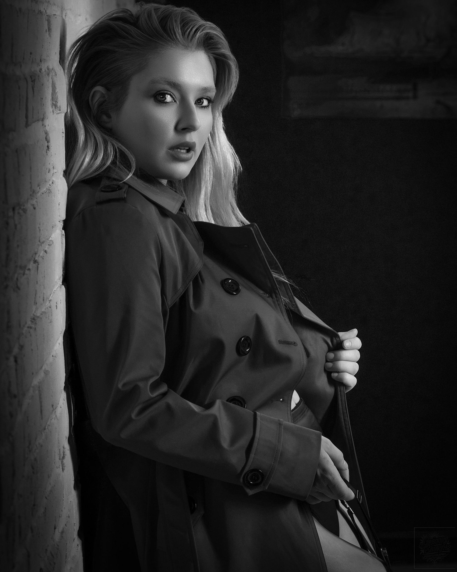 Black and white portrait of a blonde woman in a trench coat leaning against a brick wall.