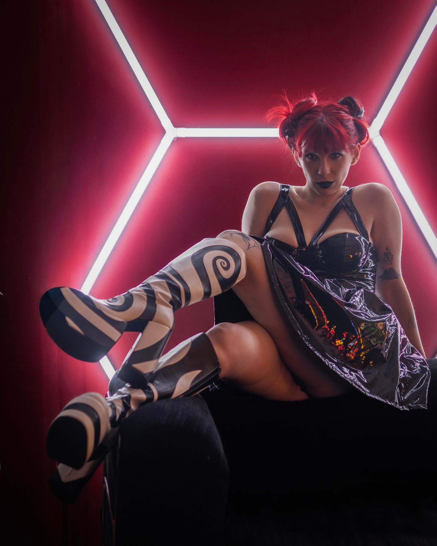 Woman in black corset dress with spiral stockings poses before glowing red hexagonal neon lights.