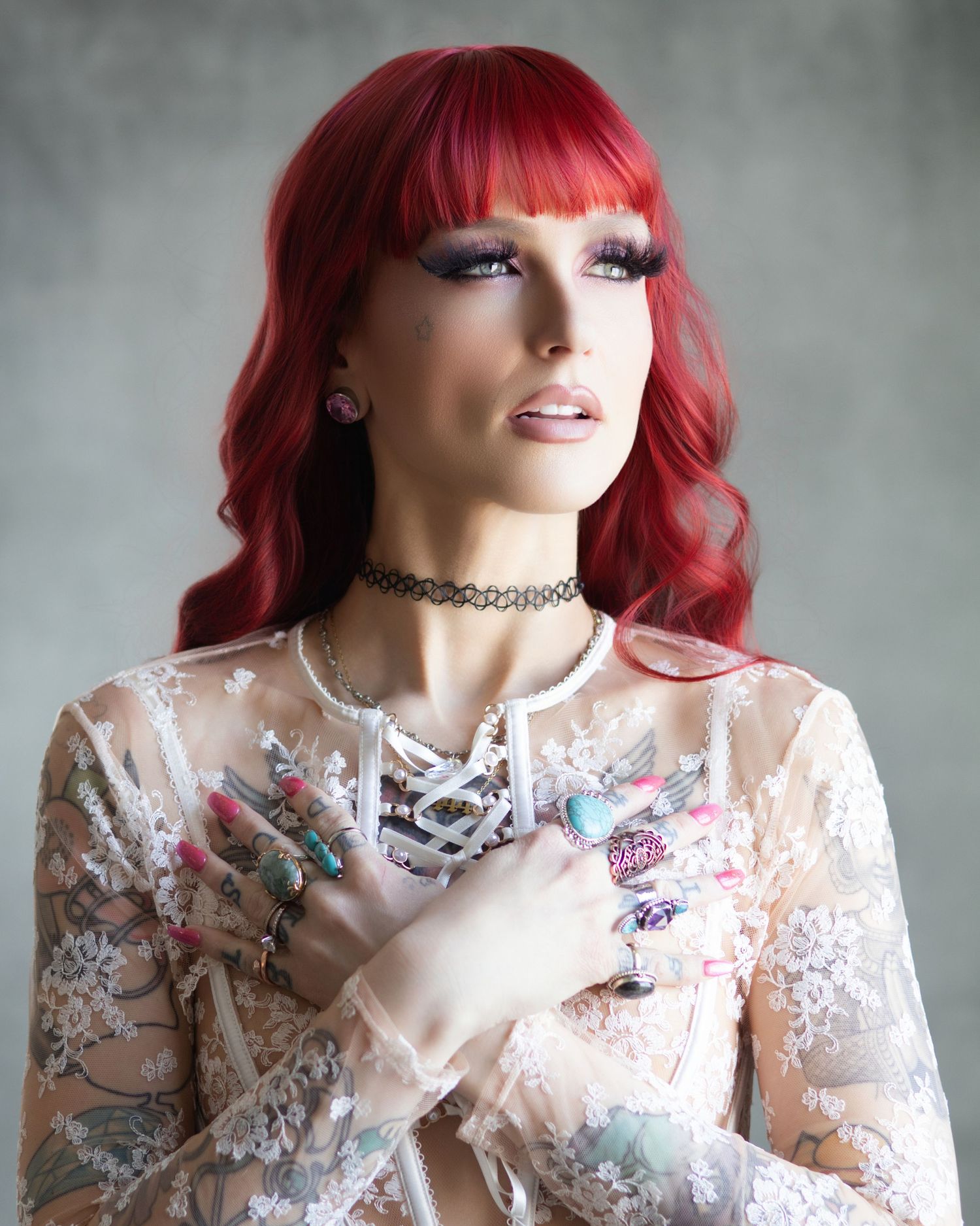 Woman with vibrant red hair, dramatic lashes, choker necklace, and floral lace top poses gracefully.