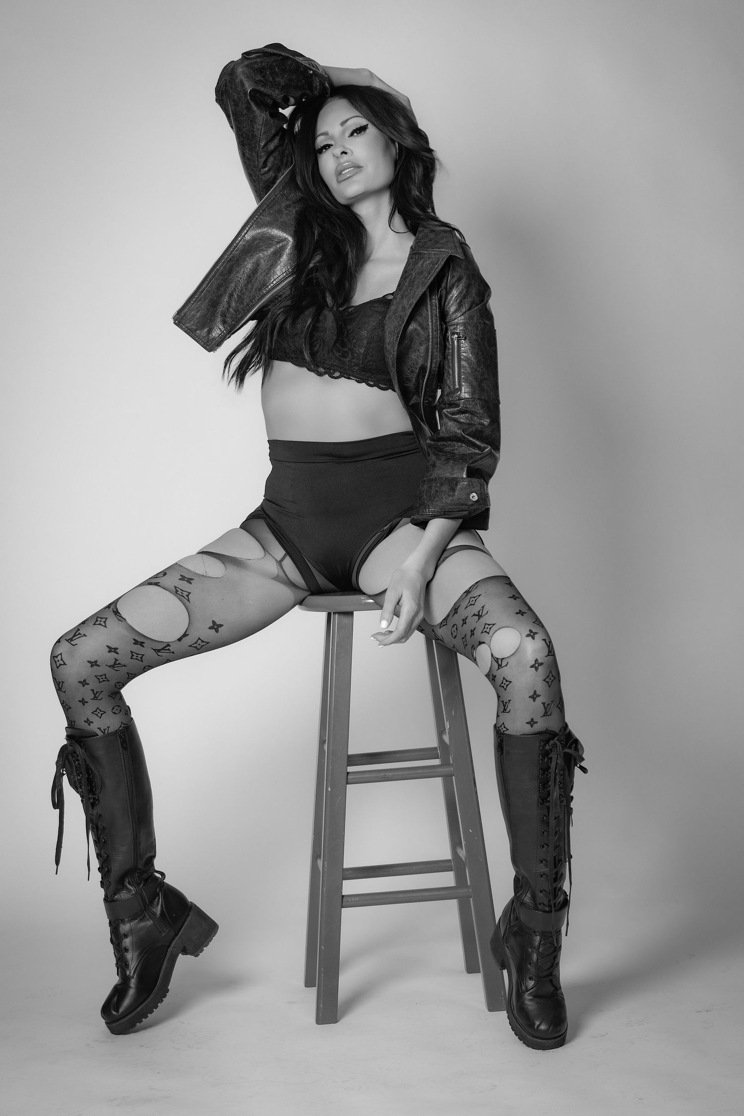 Black and white photo of a woman in leather jacket, crop top, fishnet stockings, and knee-high boots seated on a stool.