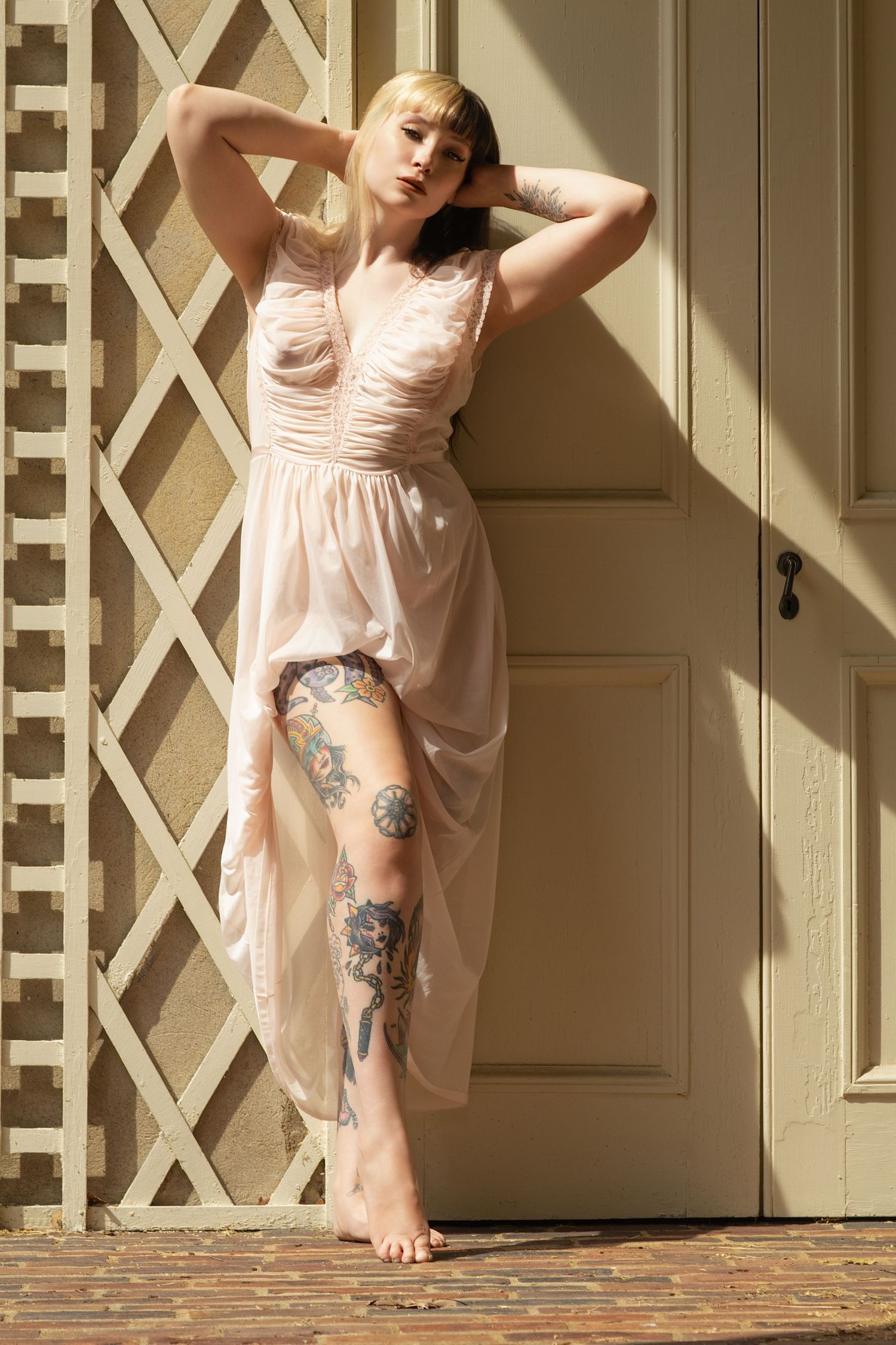 Blonde woman with leg tattoos in sheer pink dress poses barefoot against vintage white door in sunlight.