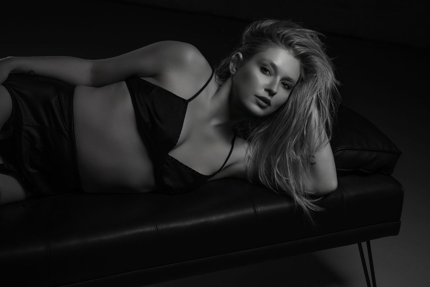 Black and white photo of a blonde woman in a black dress posing on a leather bench.