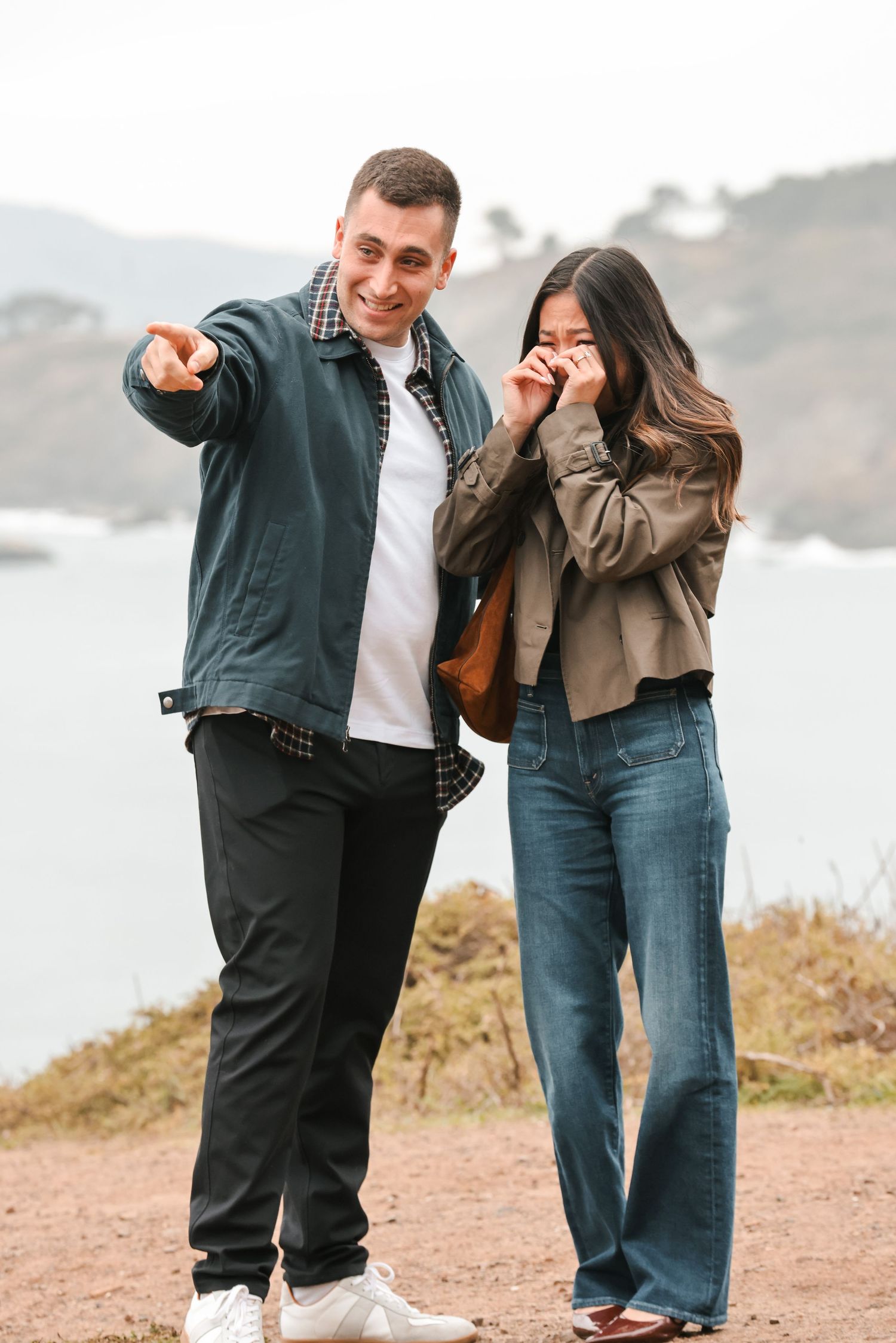 Smiling man pointing while woman laughs covering face on scenic coastal overlook on overcast day.