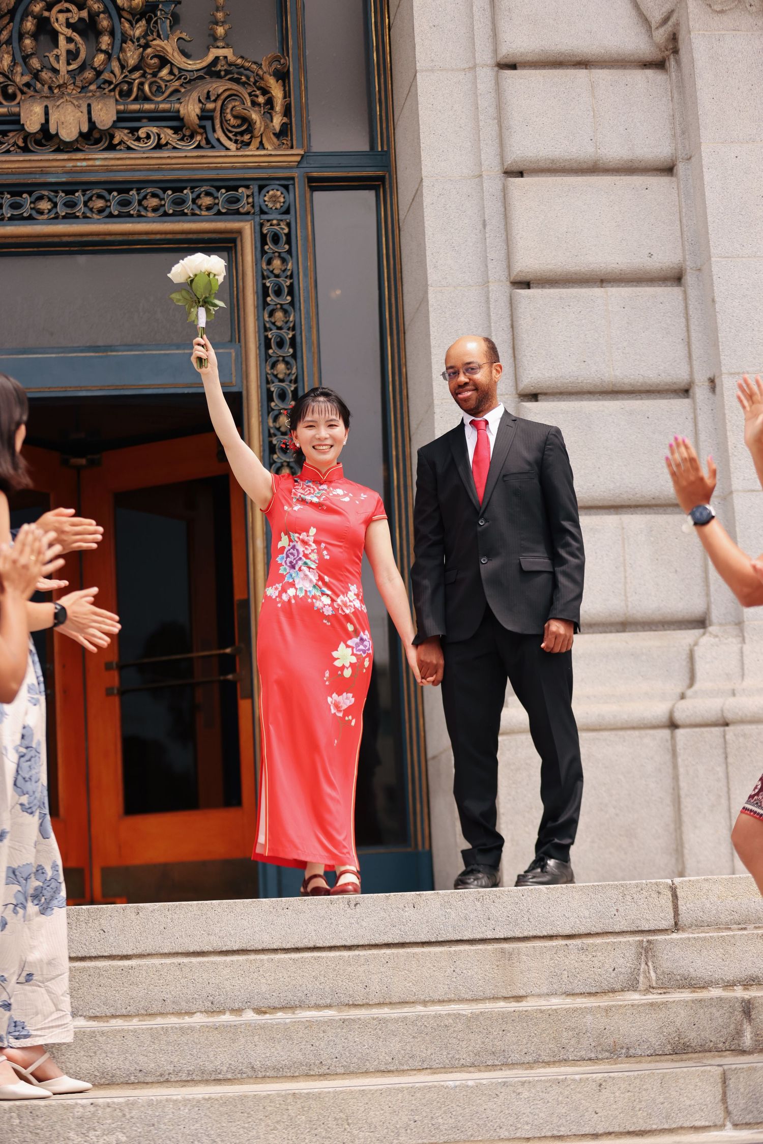 Bride in red floral qipao raises bouquet on city hall steps beside groom in black suit and red tie.