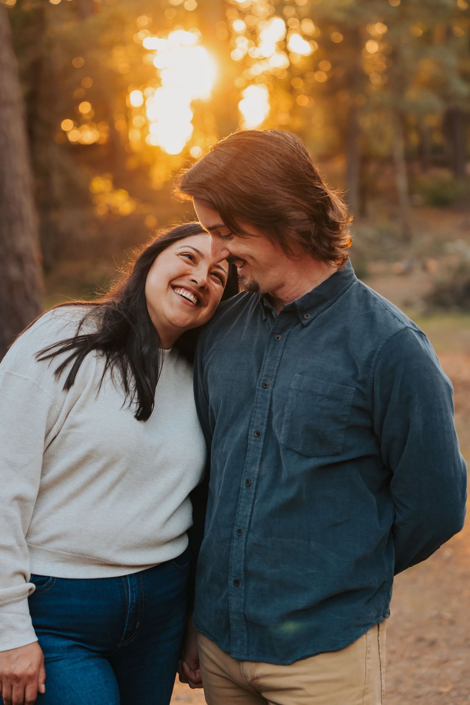 Joyful couple laughing together at golden hour in a forest, woman leans on mans shoulder.