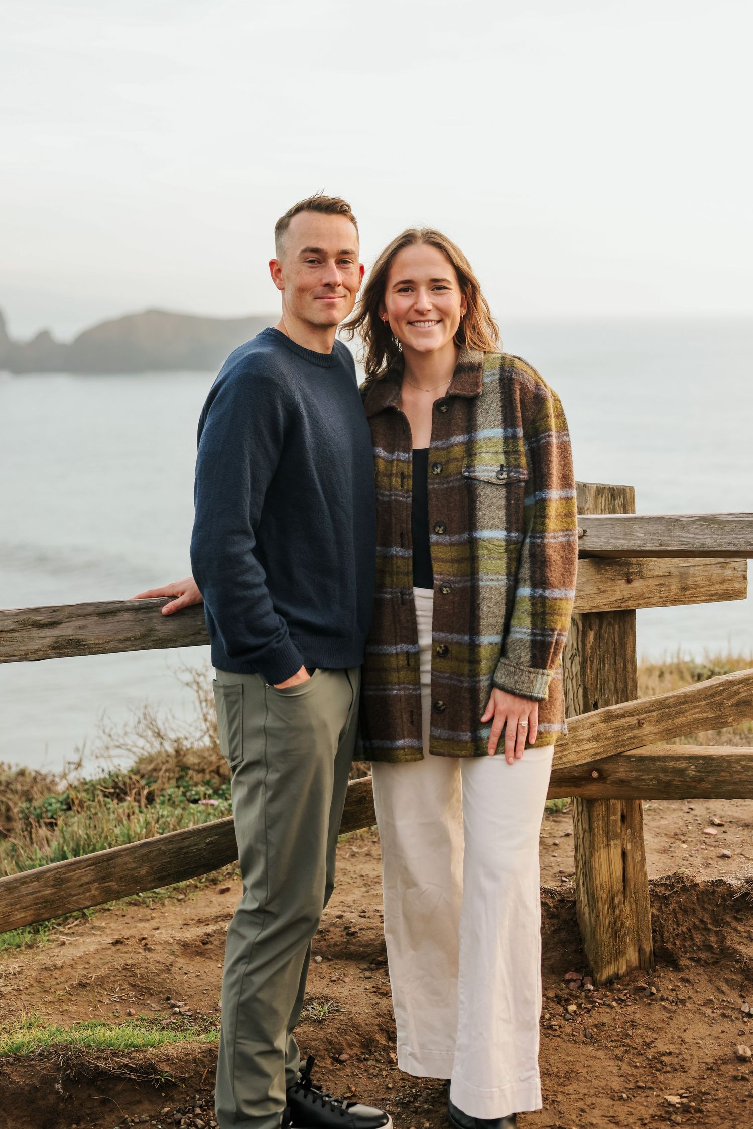 Smiling couple posing together by wooden fence with ocean view in background.