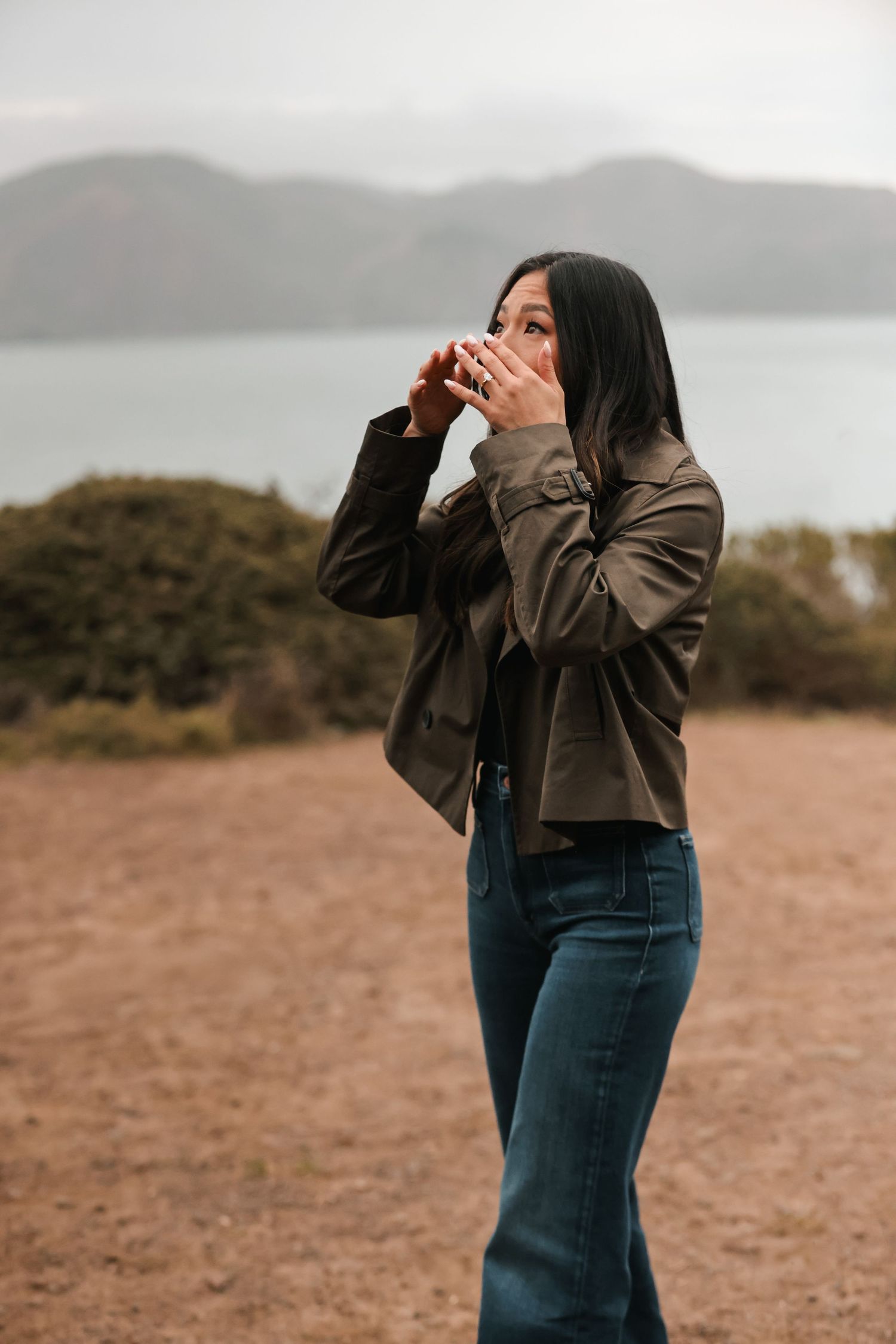 Woman covering mouth in surprise, standing outdoors near misty mountains and water.