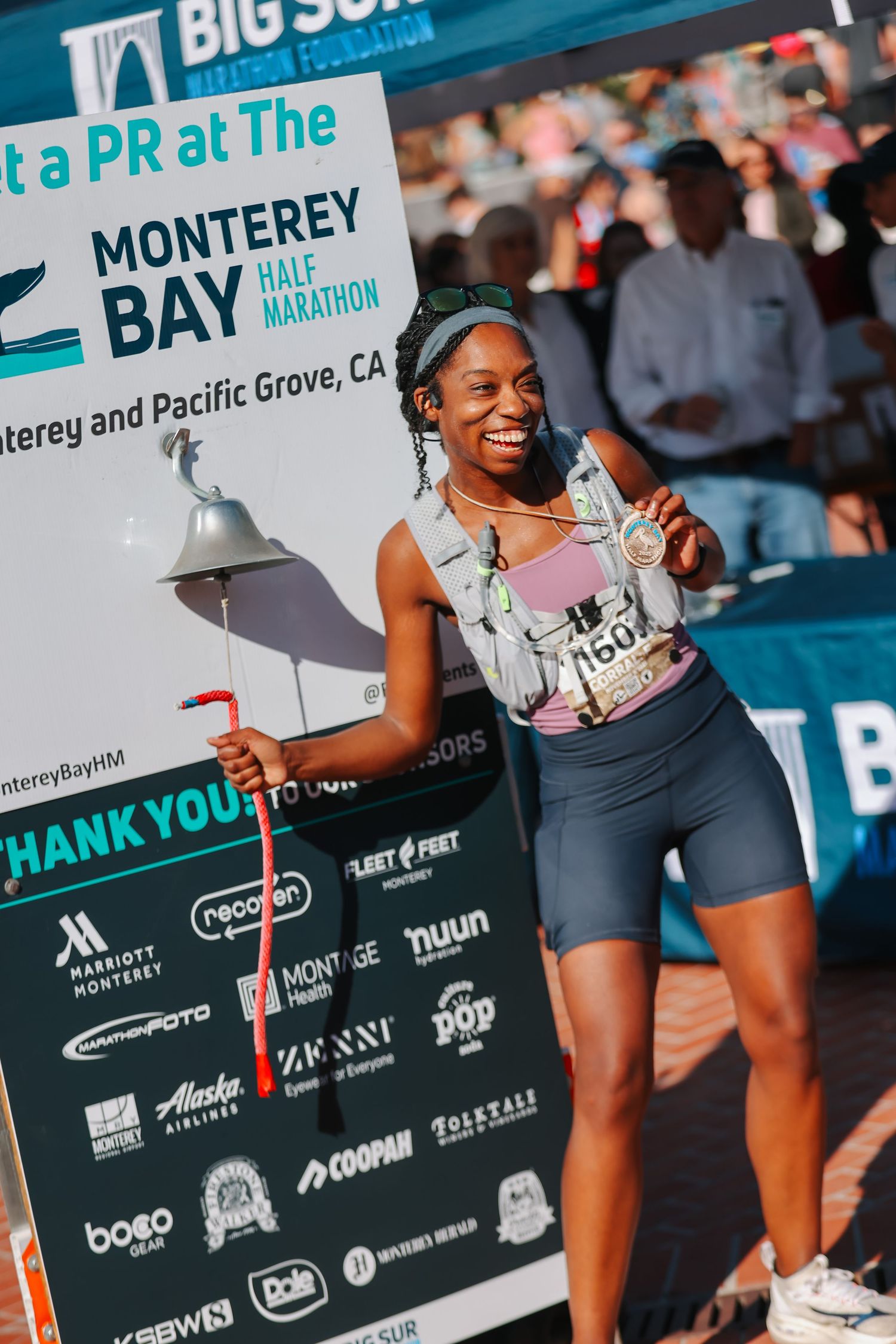 Smiling Black female runner poses with medal at Monterey Bay Half Marathon finish area holding a bell.