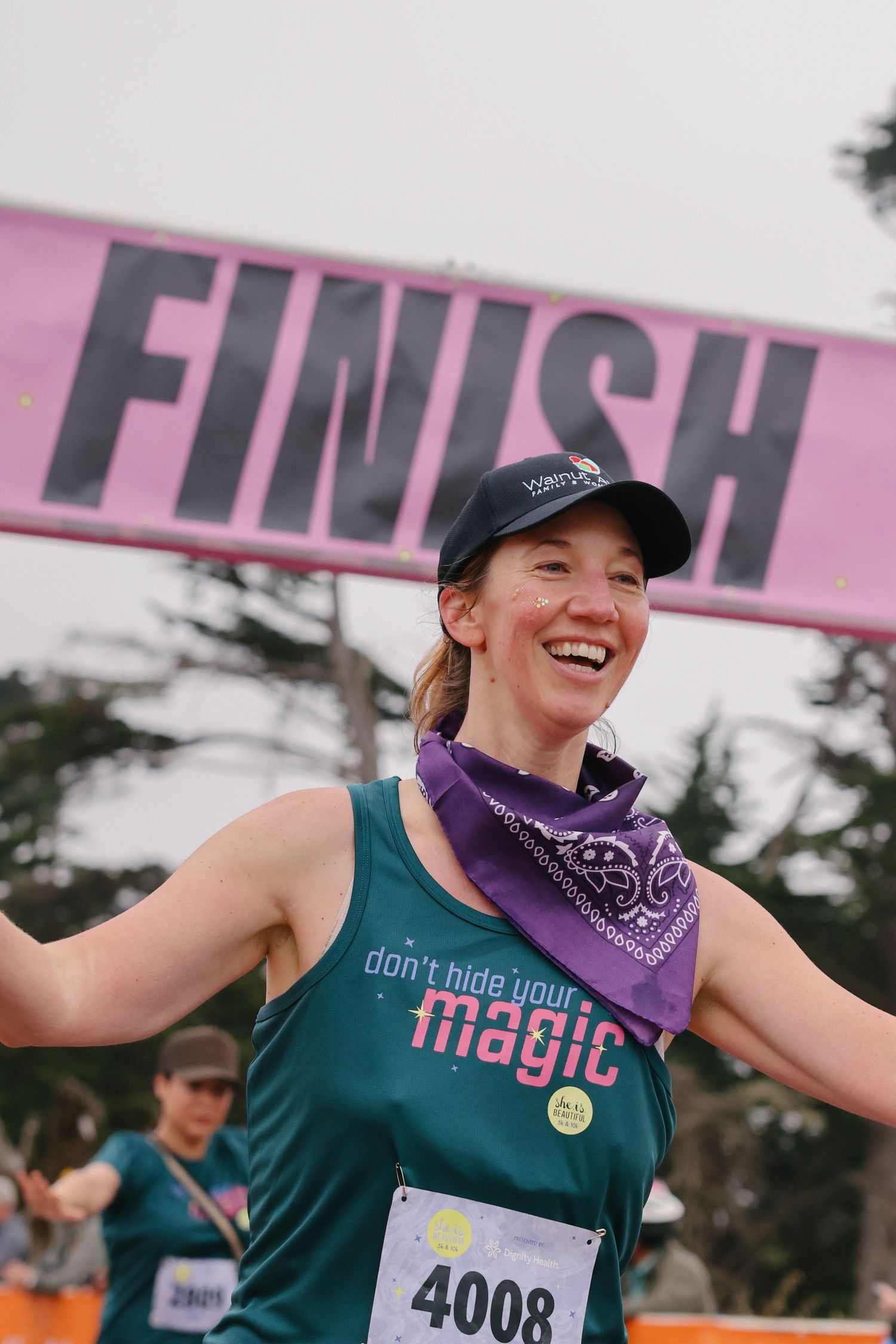 Smiling female runner crosses finish line wearing teal dont hide your magic tank top and purple bandana.