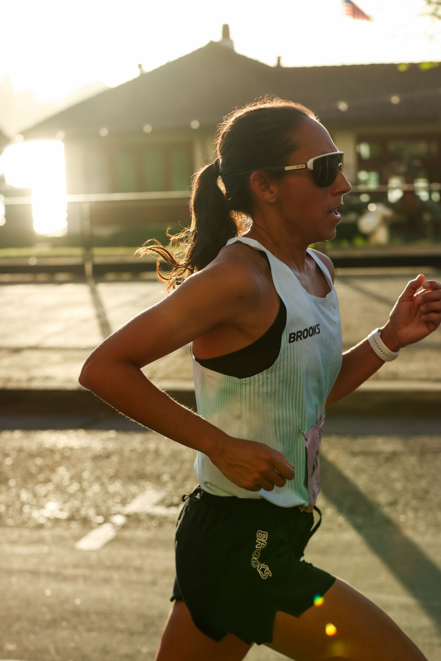 Female runner in Brooks racing vest and sunglasses runs in golden backlit sunlight during a road race.