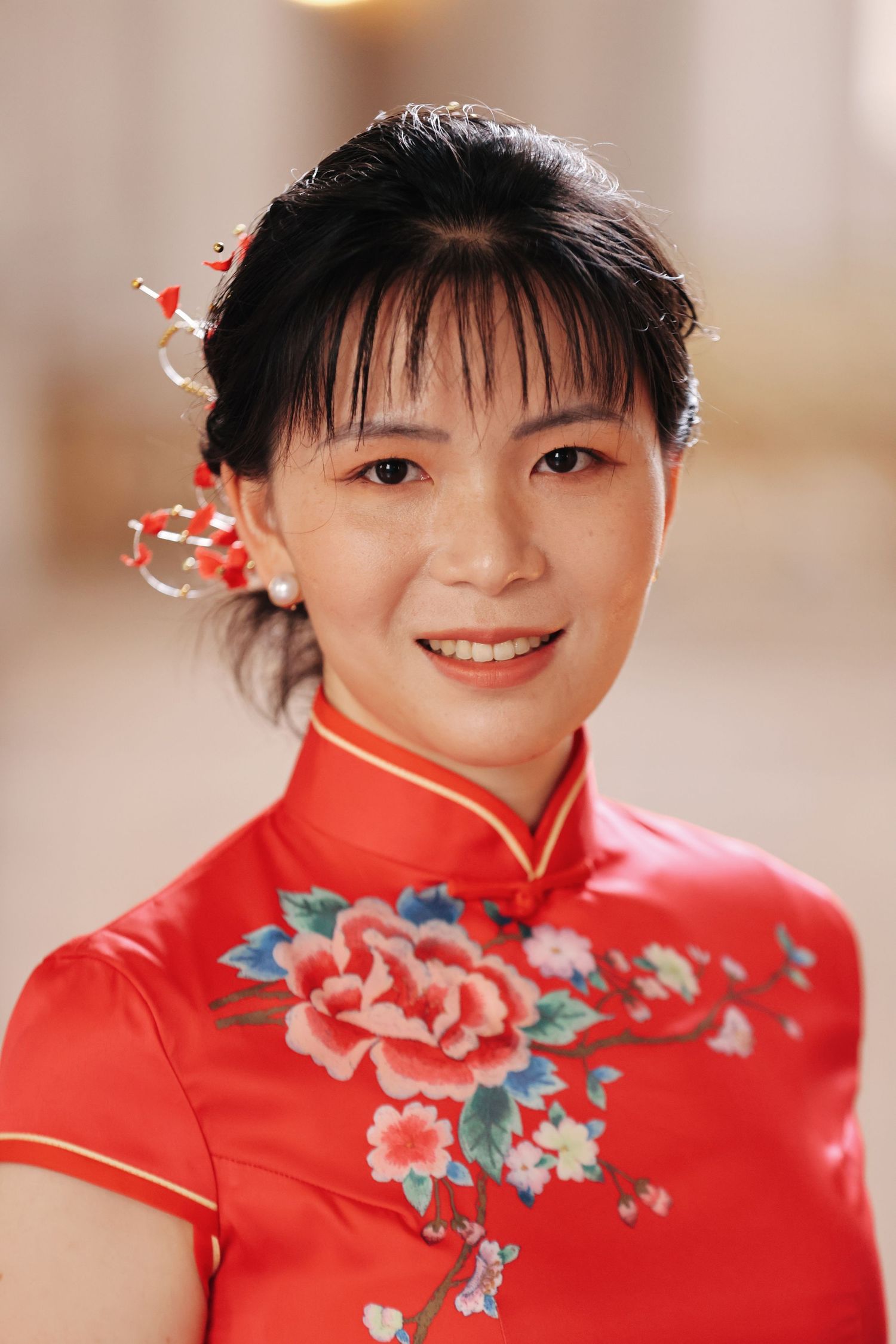 Smiling bride in a red floral cheongsam with decorative red hair accessories poses indoors.