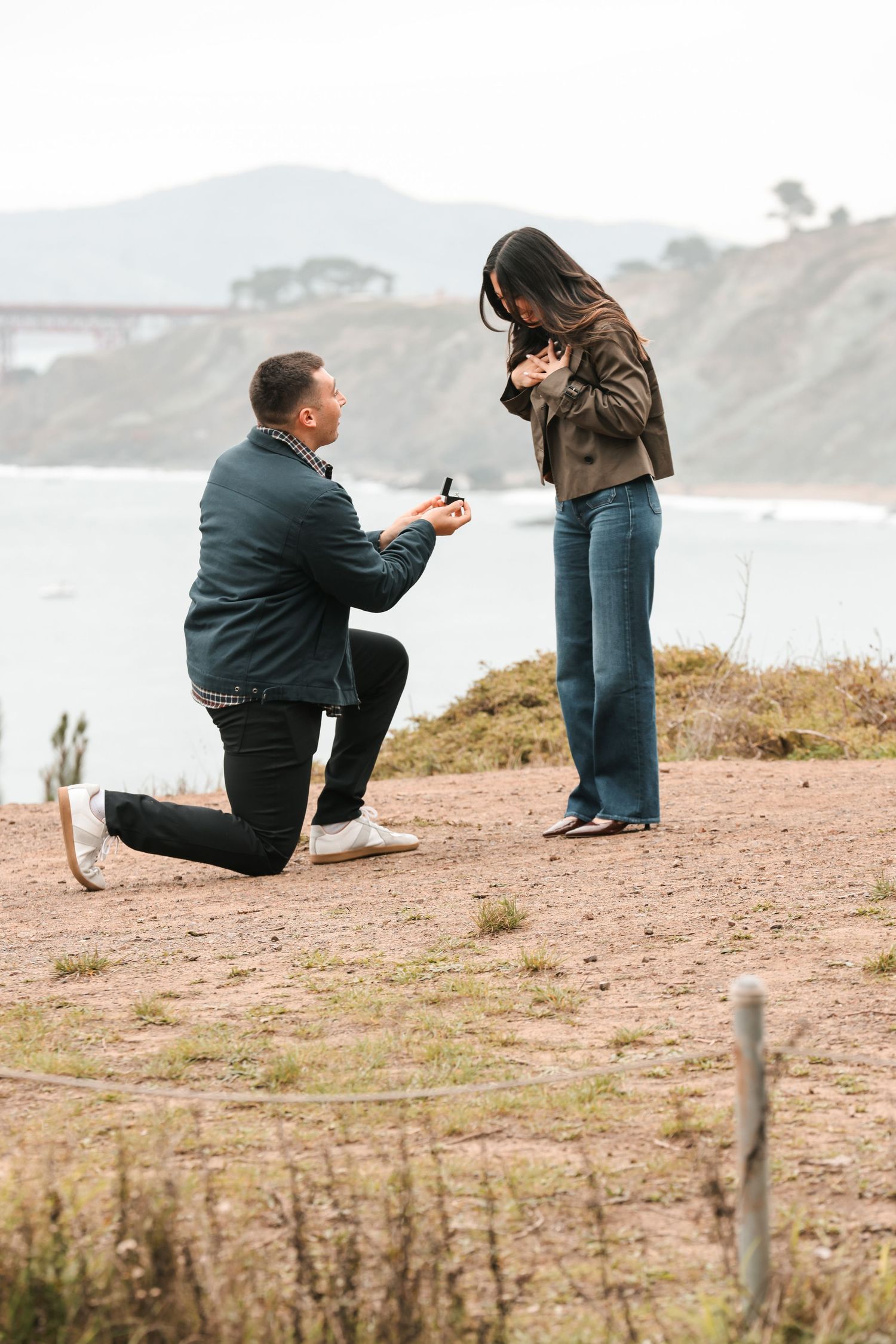 Man proposing on one knee with ring box to woman outdoors near coastal cliffs with Golden Gate Bridge in background.
