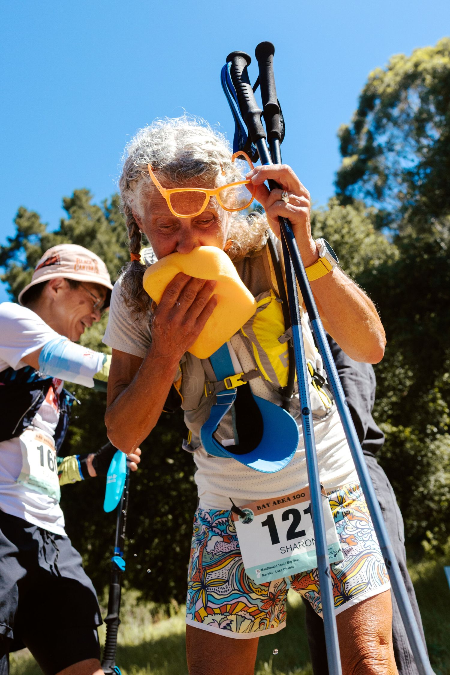 Elderly trail runner with trekking poles drinking from yellow bottle at outdoor race event on sunny day.