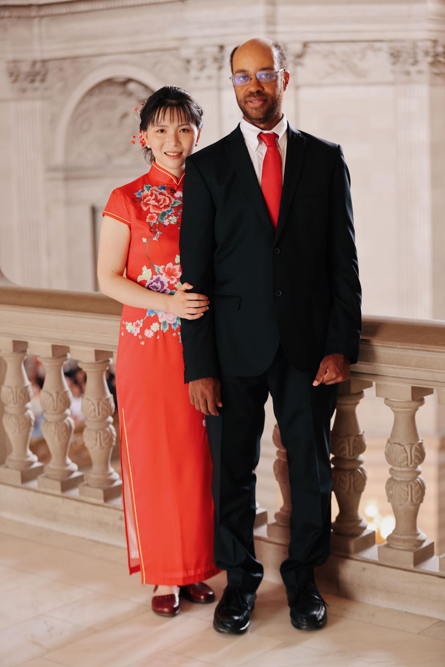 Interracial wedding couple posing on elegant marble staircase, bride in red qipao and groom in black suit.